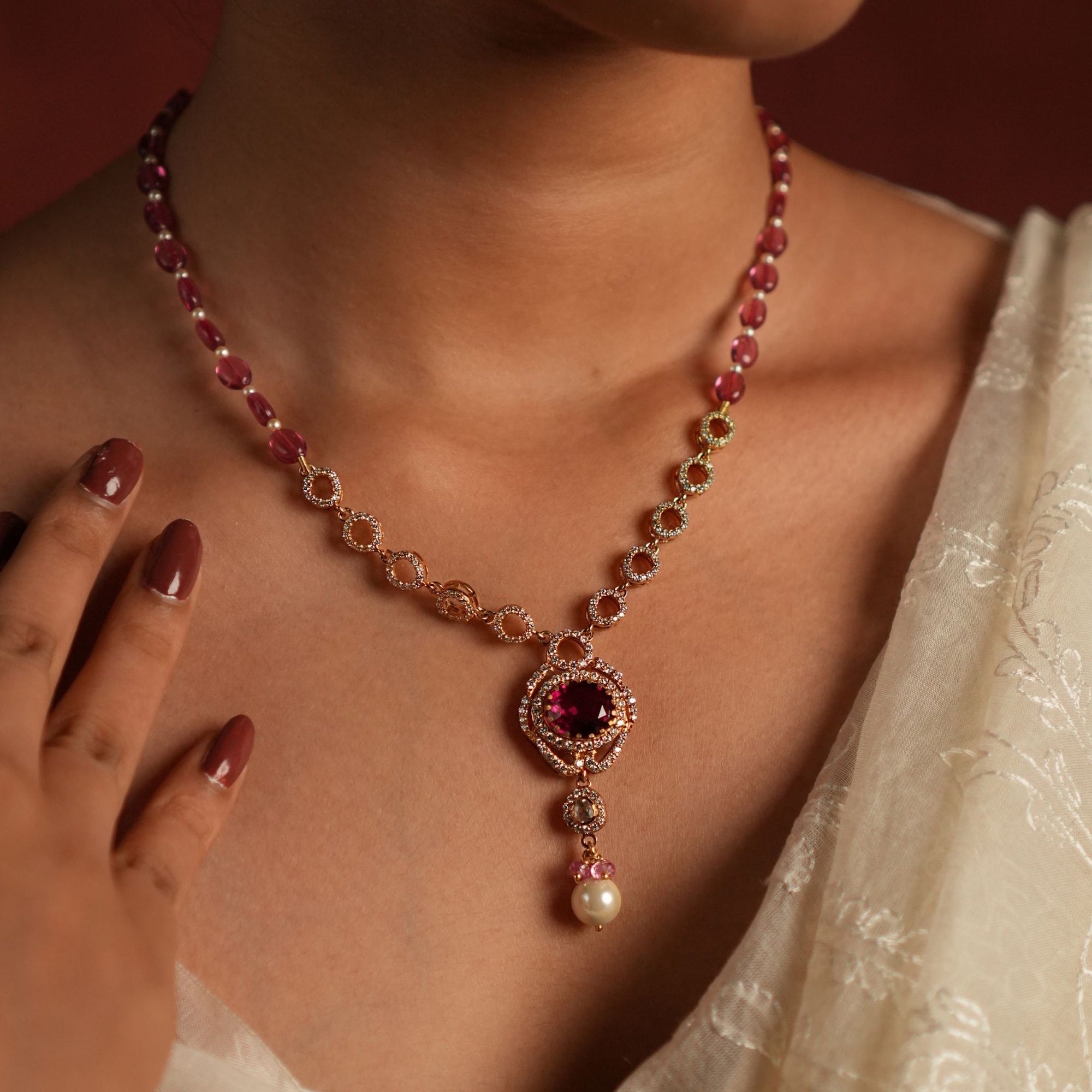 Model wearing an elegant gold-toned necklace with ruby-coloured beads, a diamond-accented circular pendant, and a pearl drop, styled with an ivory saree.
