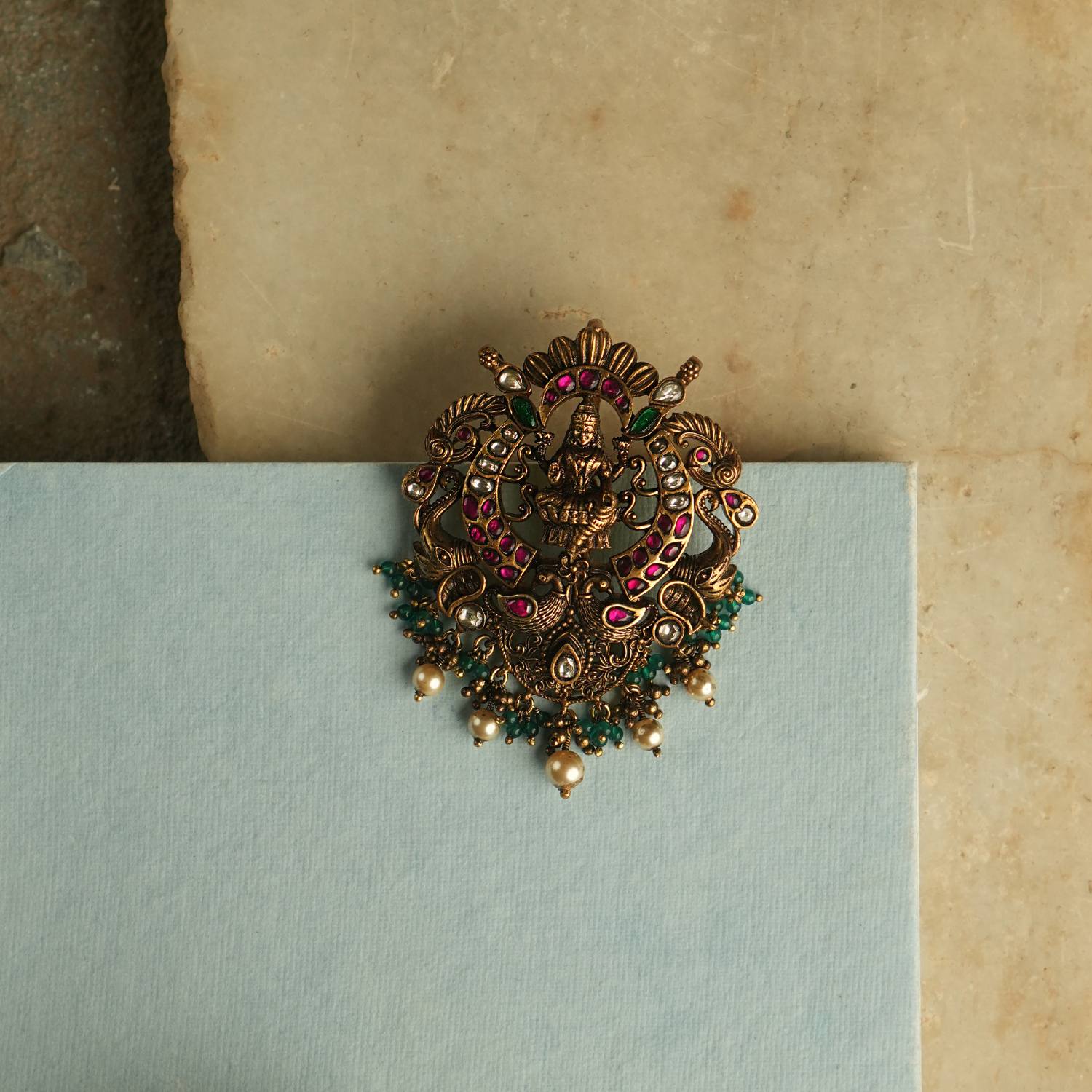 A fusion of elegance, heritage, and contemporary style, this antique-plated pendant features red, green, and white Kundan stones, with a divine goddess at its center. Delicate white and green beads cascade as droplets, adding timeless charm to this exquisite piece.