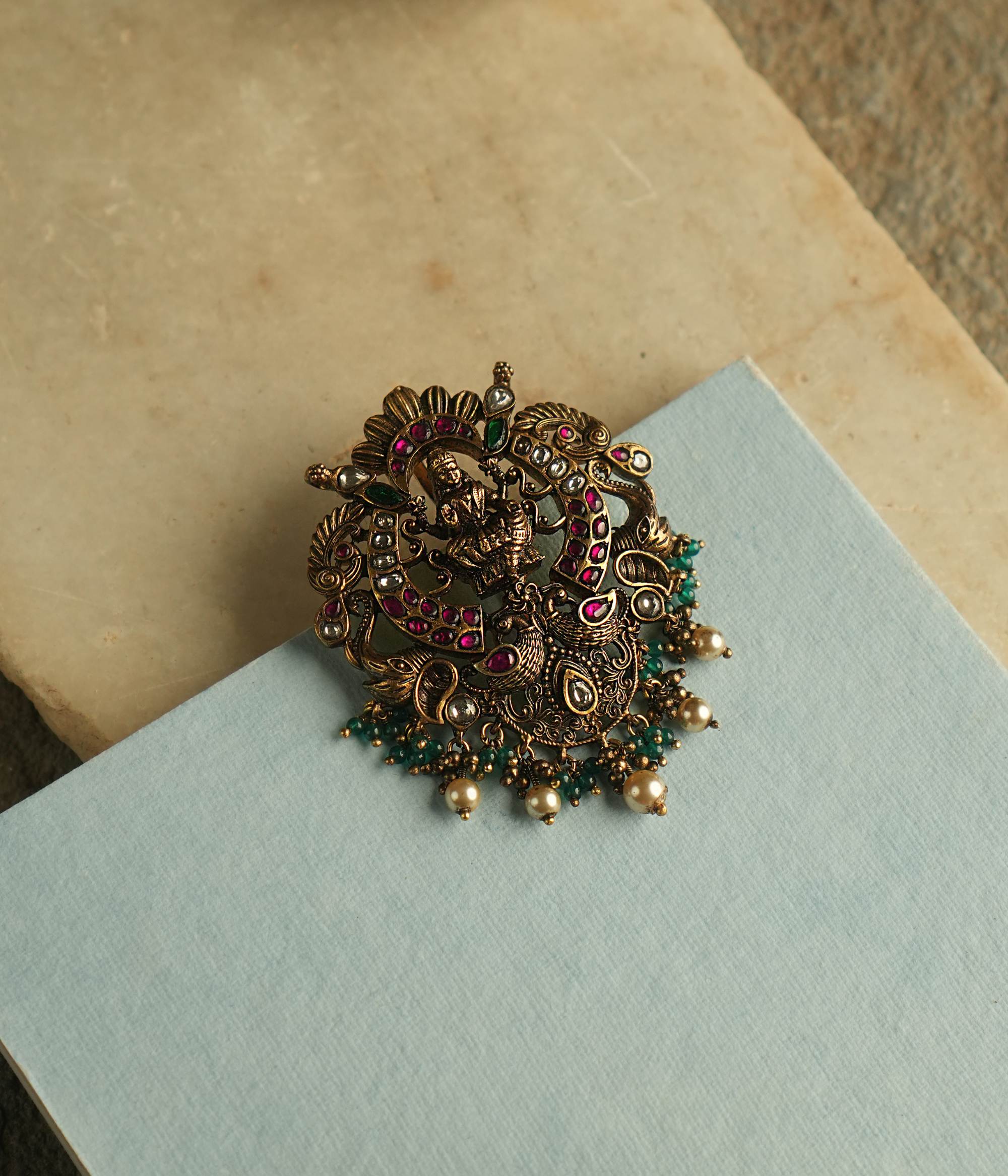A fusion of elegance, heritage, and contemporary style, this antique-plated pendant features red, green, and white Kundan stones, with a divine goddess at its center. Delicate white and green beads cascade as droplets, adding timeless charm to this exquisite piece.