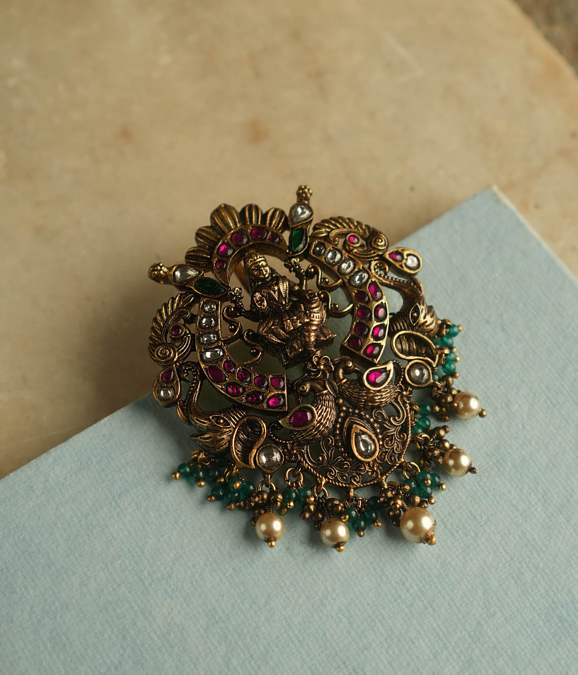 A fusion of elegance, heritage, and contemporary style, this antique-plated pendant features red, green, and white Kundan stones, with a divine goddess at its center. Delicate white and green beads cascade as droplets, adding timeless charm to this exquisite piece.