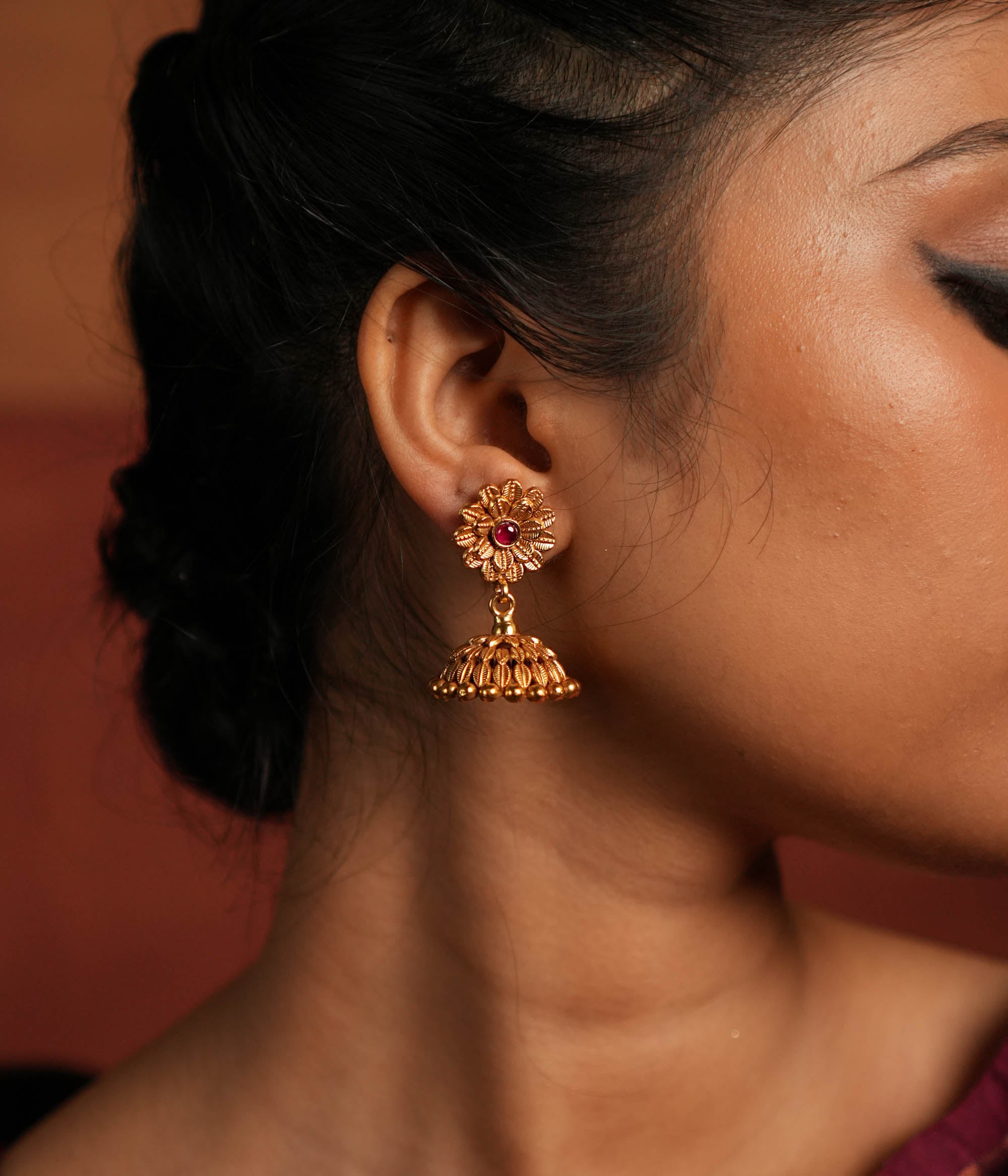 Exquisite jhumka earrings featuring a floral design engraved with intricate tiny floral details. The centerpiece showcases a striking red Kundan stone, enhancing the elegance of the design. The jhumka structure adds a traditional touch, making it a timeless statement piece.