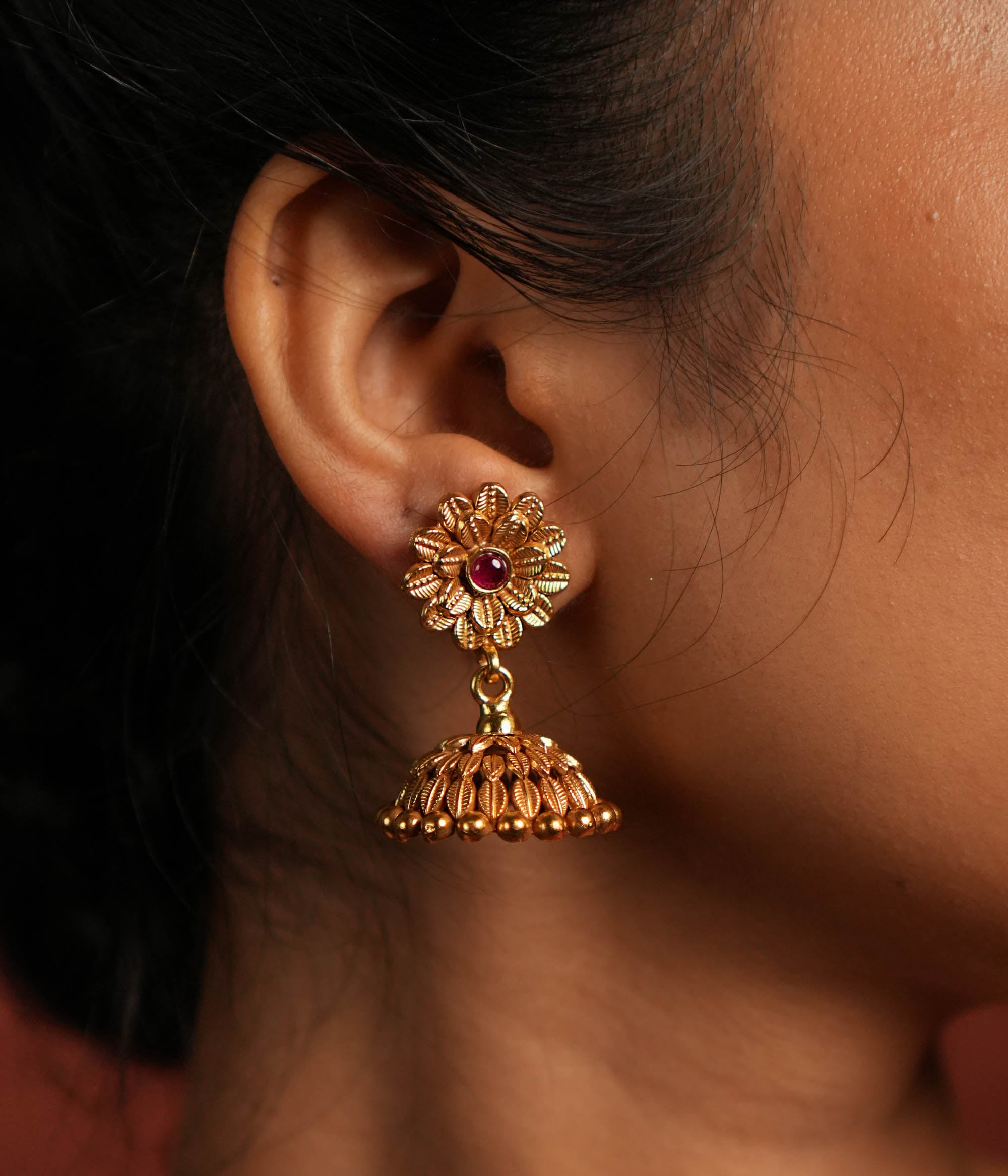 Exquisite jhumka earrings featuring a floral design engraved with intricate tiny floral details. The centerpiece showcases a striking red Kundan stone, enhancing the elegance of the design. The jhumka structure adds a traditional touch, making it a timeless statement piece.