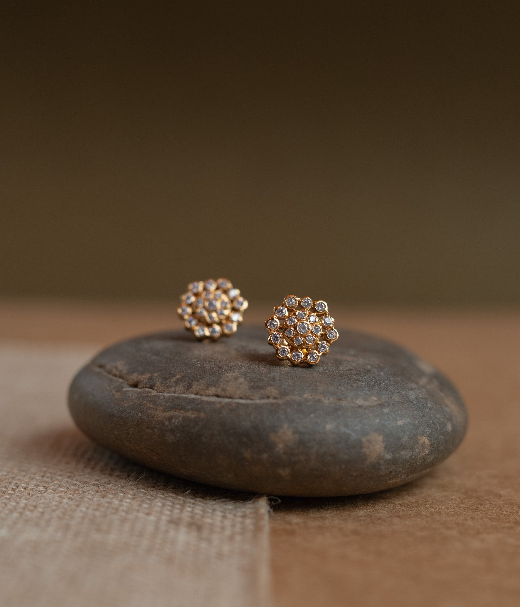 Gold floral stud earrings covered in clear Swarovski stones, displayed on a textured fabric.