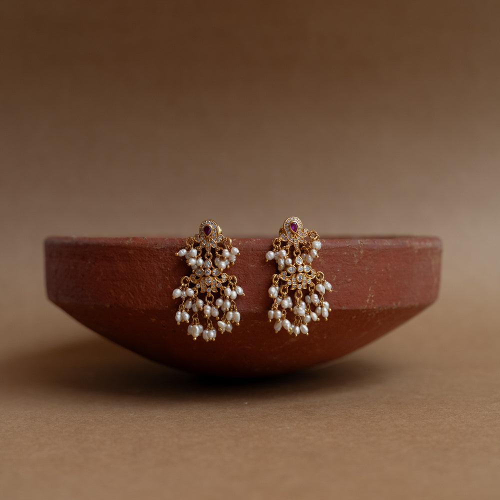 Gold drop earrings with Swarovski stones, a red cut stone, and multiple dangling pearl clusters, displayed on a brass tray.