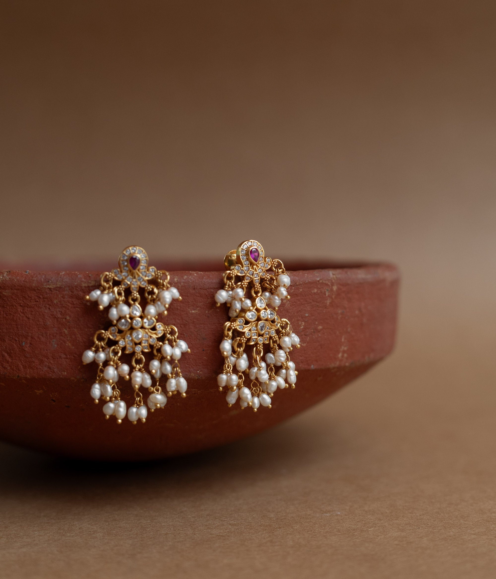 Gold drop earrings with Swarovski stones, a red cut stone, and multiple dangling pearl clusters, displayed on a brass tray.