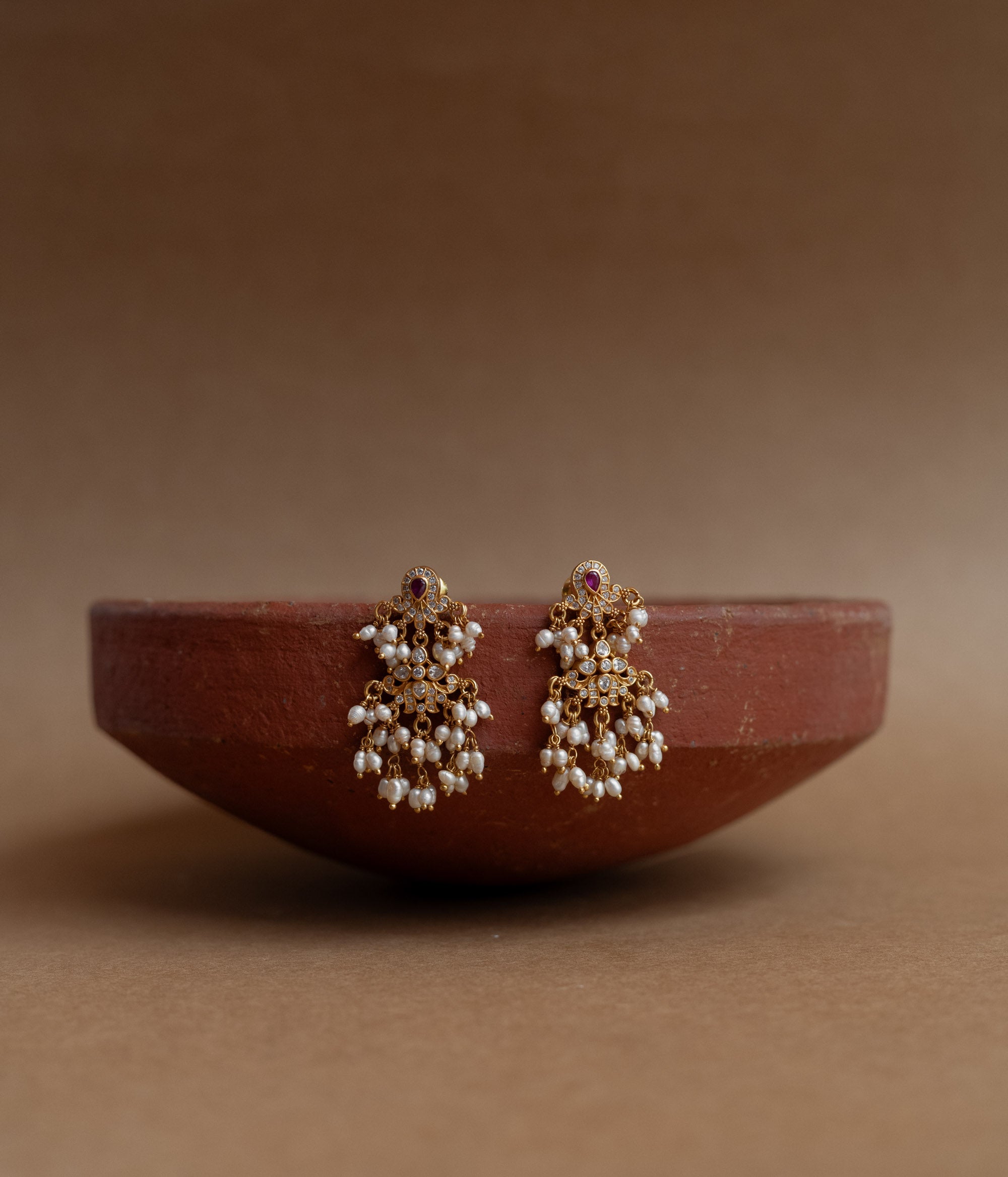 Gold drop earrings with Swarovski stones, a red cut stone, and multiple dangling pearl clusters, displayed on a brass tray.