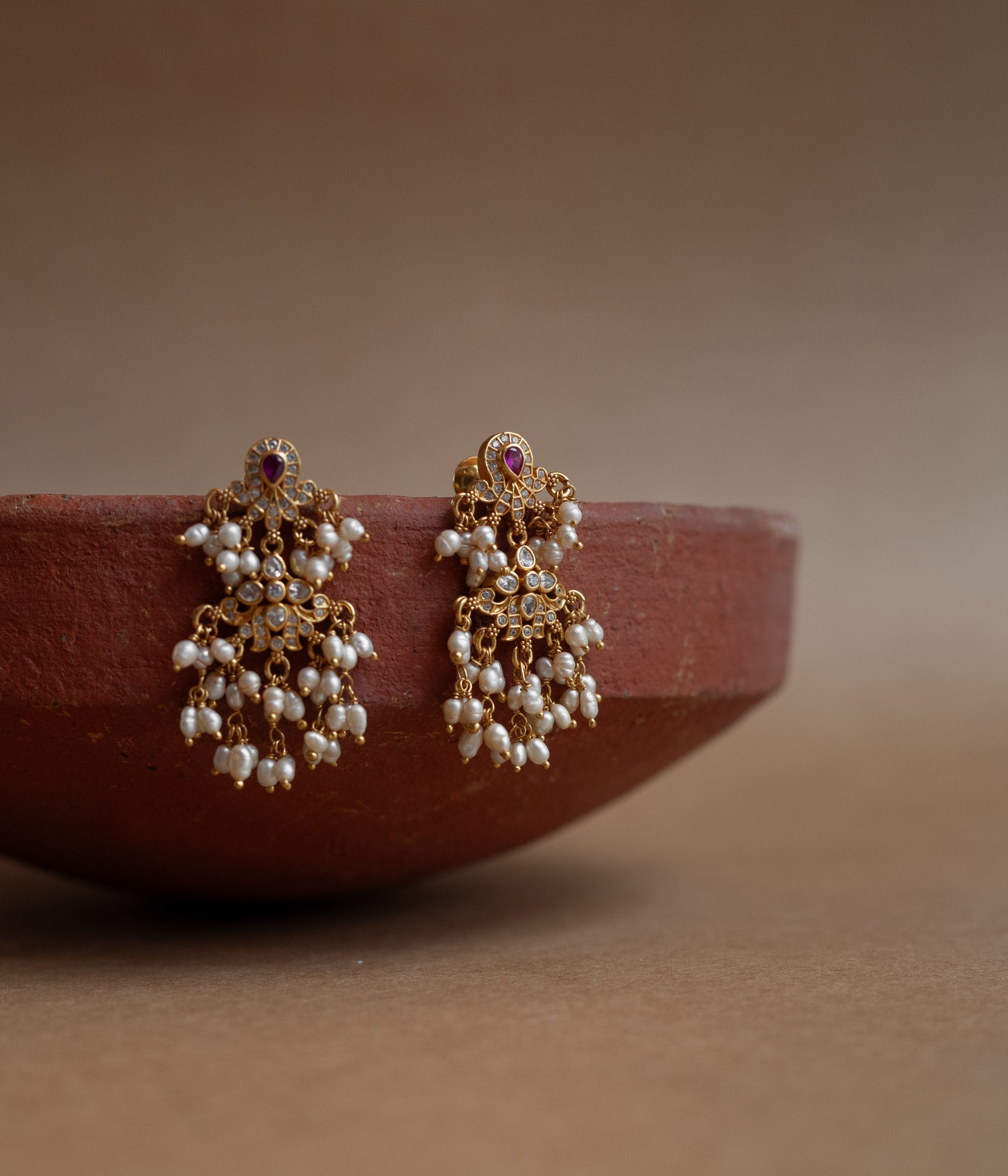 Gold drop earrings with Swarovski stones, a red cut stone, and multiple dangling pearl clusters, displayed on a brass tray.