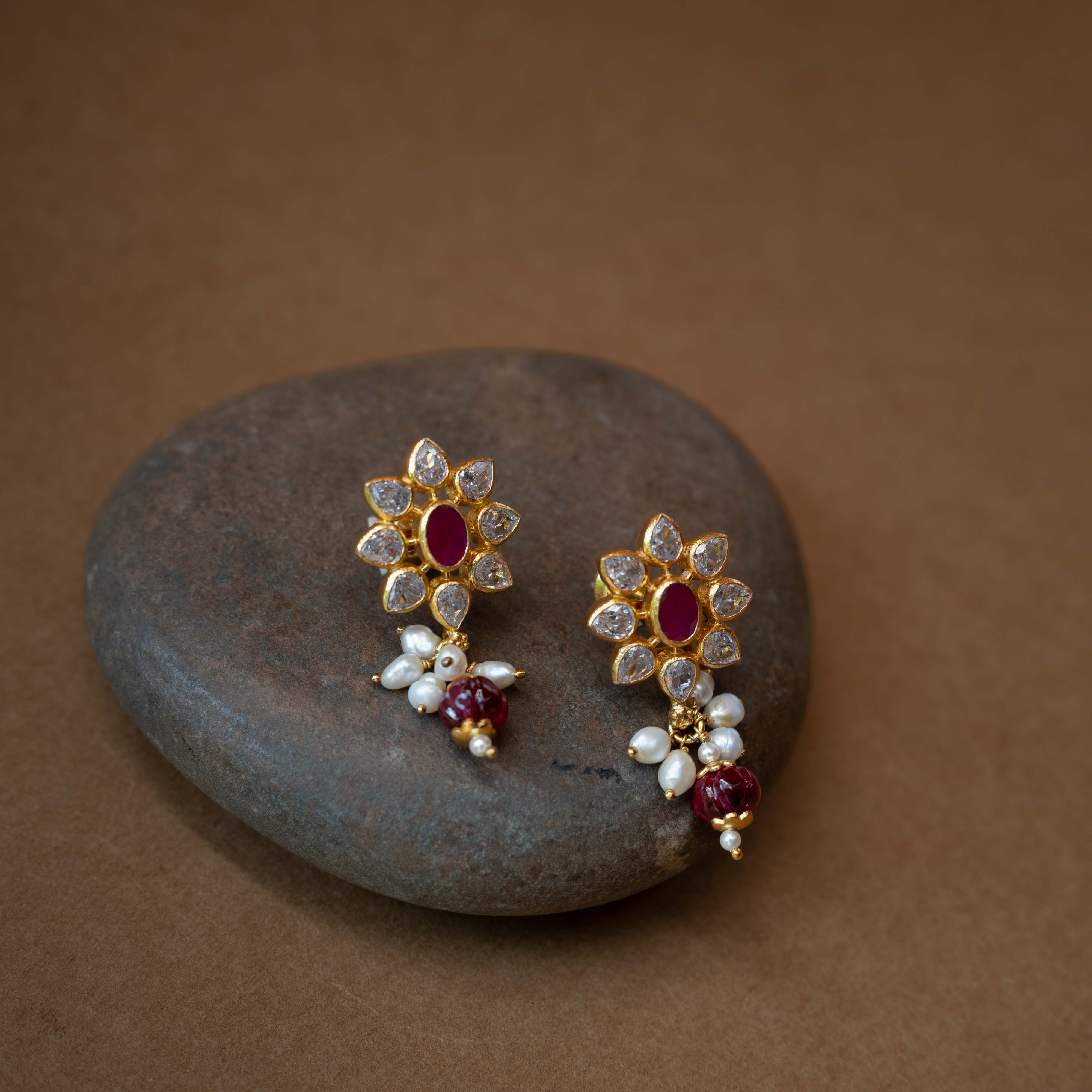 A pair of intricate floral earrings with a central red stone, surrounded by clear stones, and dangling pearls.
