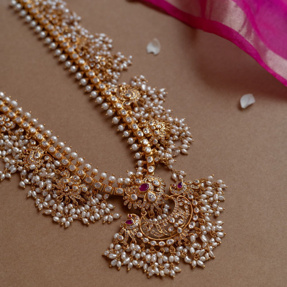 A long gold necklace adorned with numerous small pearls. It features a central pendant with clear and red stones.