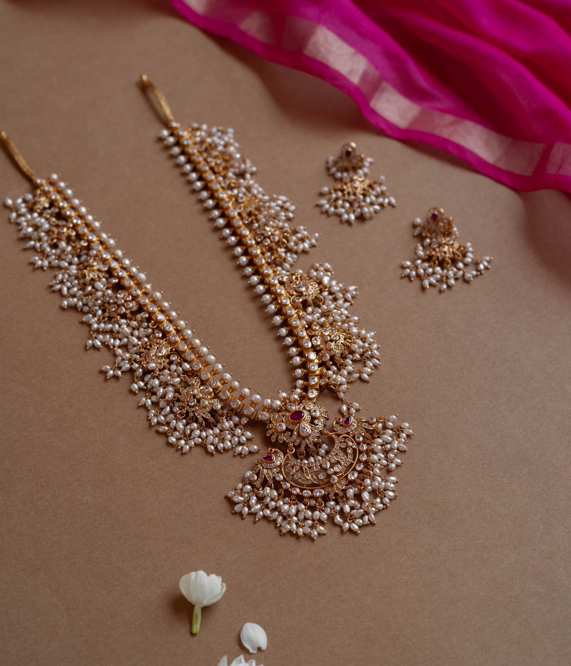A long gold necklace adorned with numerous small pearls. It features a central pendant with clear and red stones.