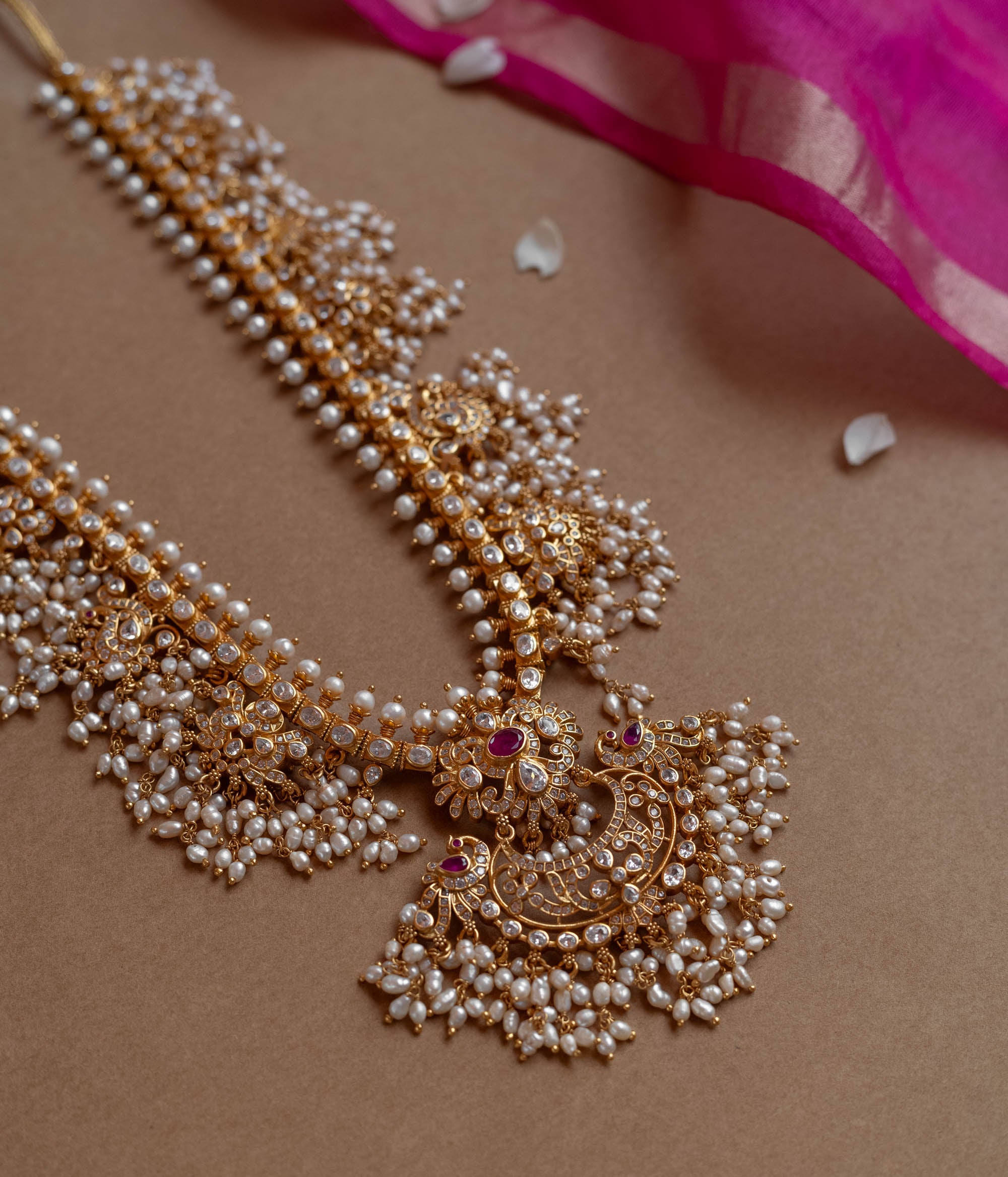 A long gold necklace adorned with numerous small pearls. It features a central pendant with clear and red stones.
