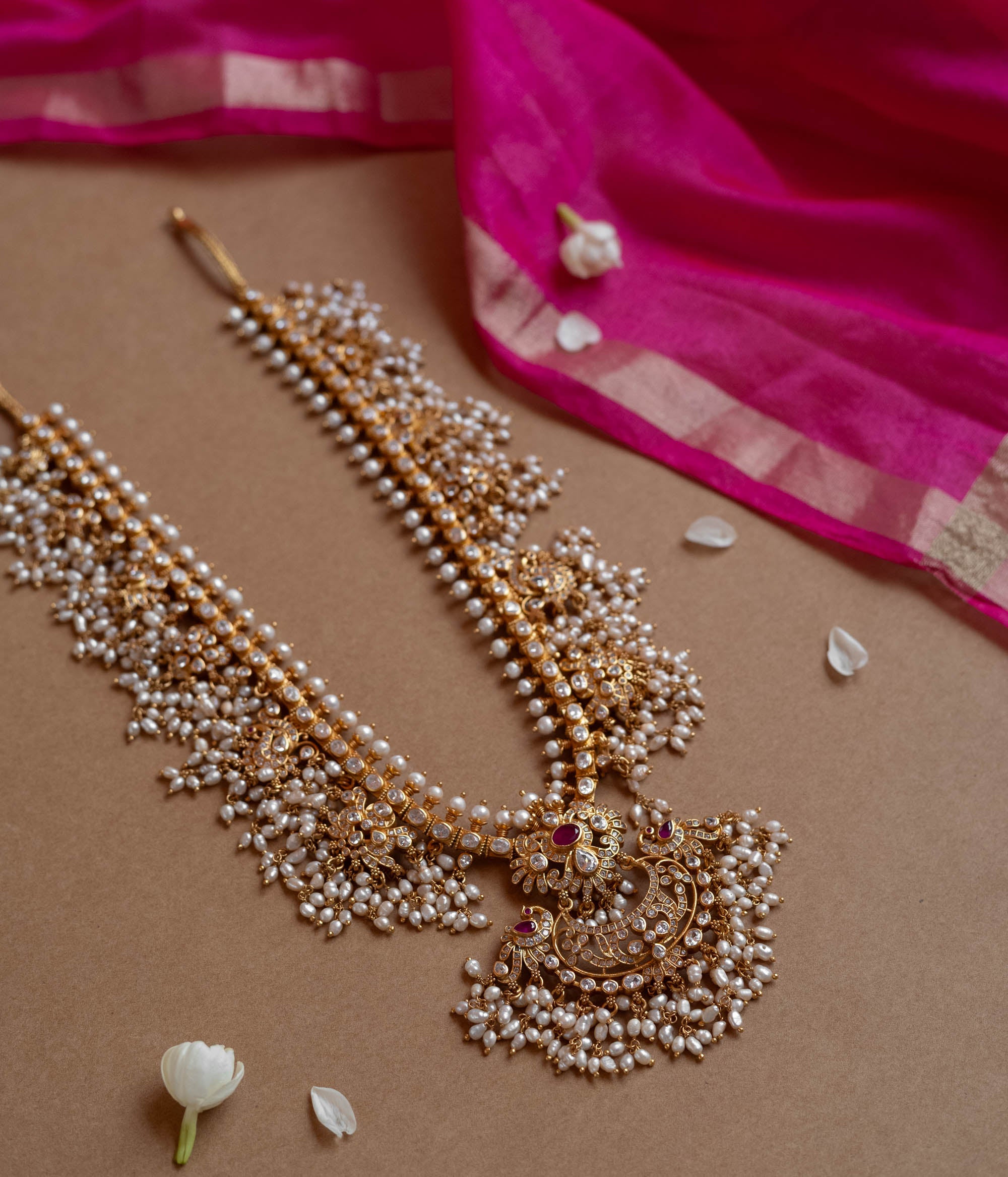 A long gold necklace adorned with numerous small pearls. It features a central pendant with clear and red stones.