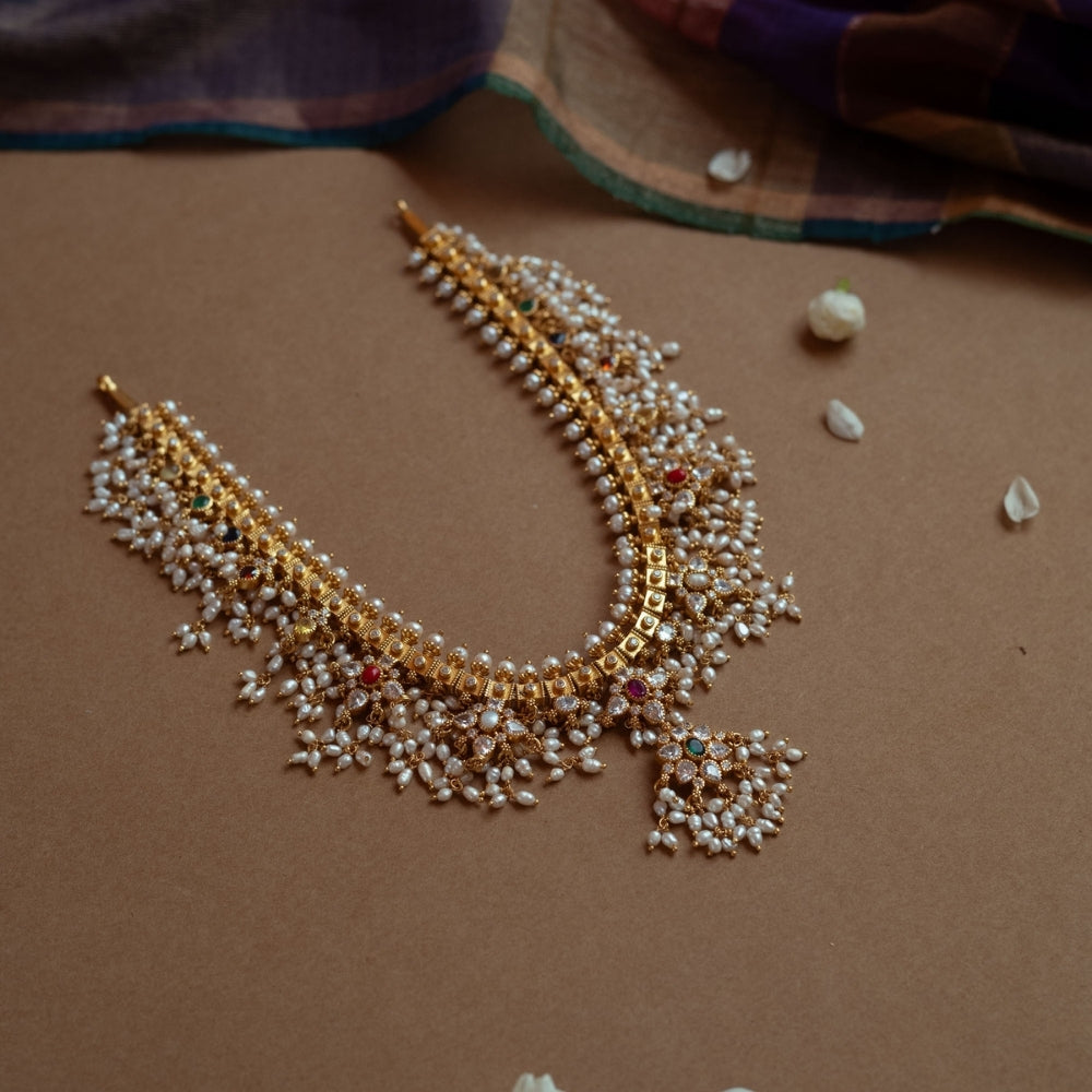A gold necklace with a dense cascade of small white pearls. It features embedded clear, red, and green stones, and rests on a brown surface with white flower petals.