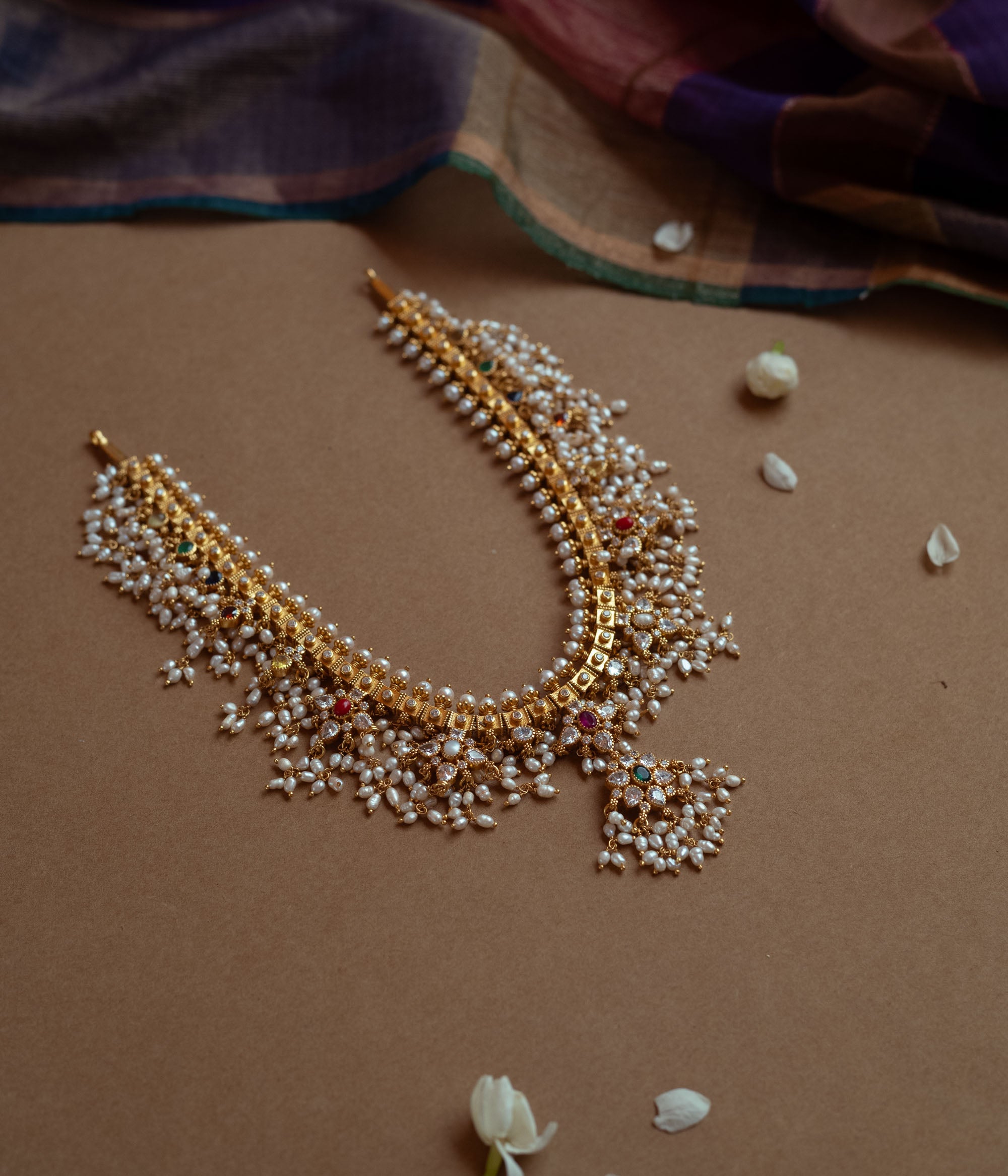 A gold necklace with a dense cascade of small white pearls. It features embedded clear, red, and green stones, and rests on a brown surface with white flower petals.