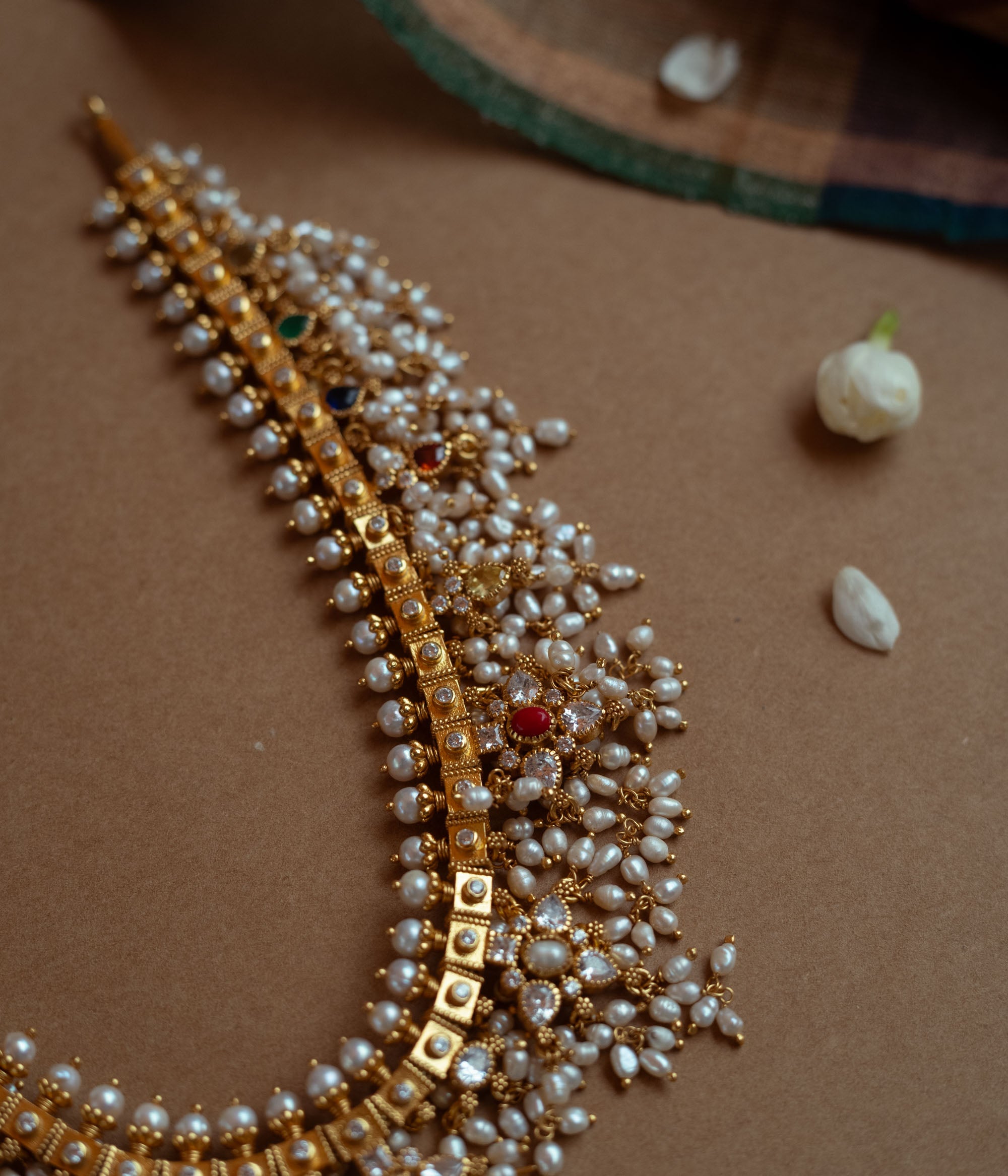 A gold necklace with a dense cascade of small white pearls. It features embedded clear, red, and green stones, and rests on a brown surface with white flower petals.