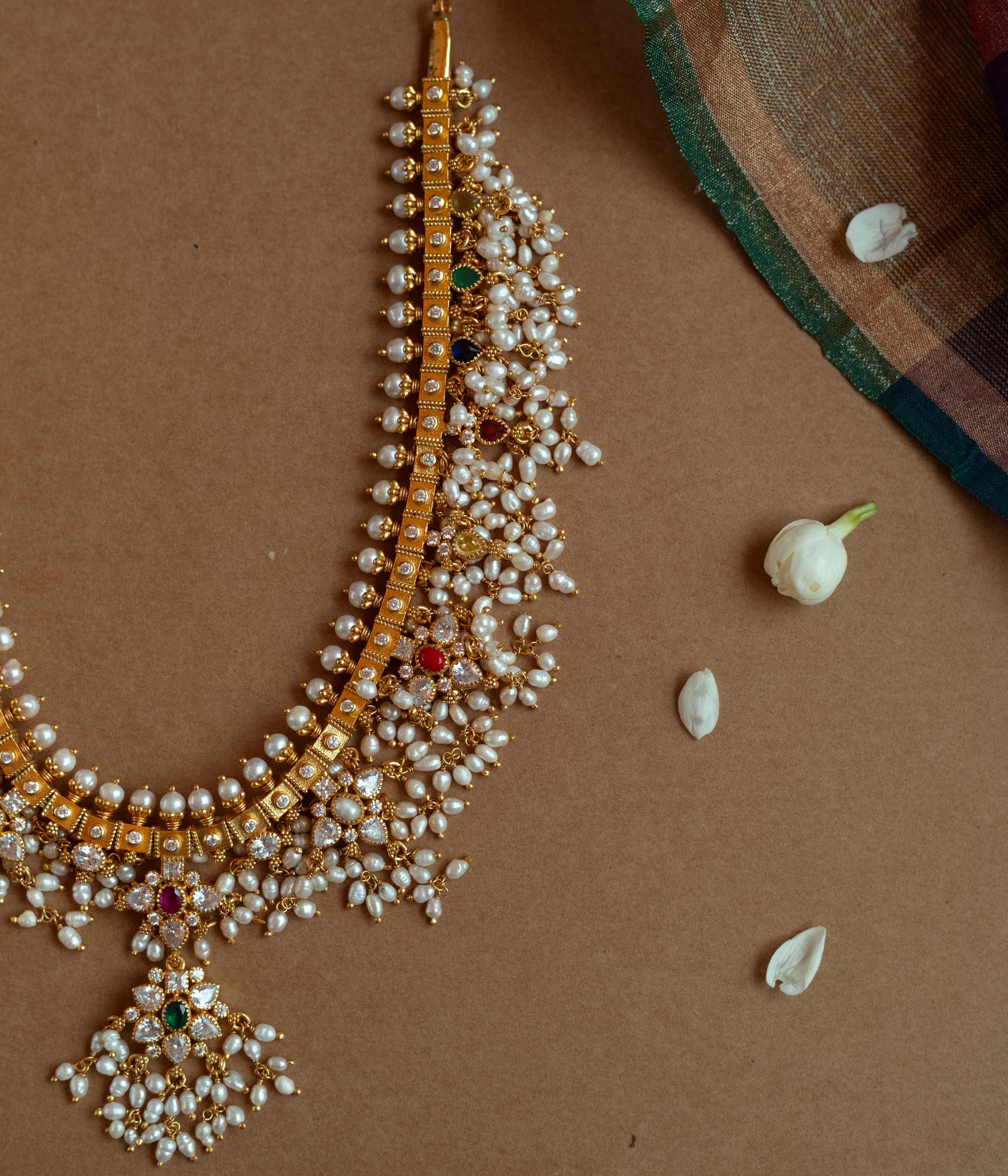 A gold necklace with a dense cascade of small white pearls. It features embedded clear, red, and green stones, and rests on a brown surface with white flower petals.