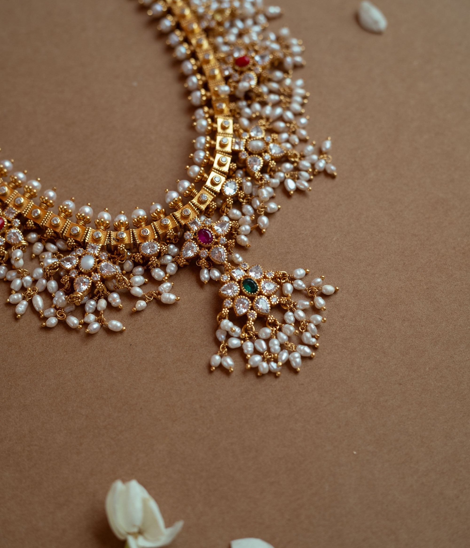 A gold necklace with a dense cascade of small white pearls. It features embedded clear, red, and green stones, and rests on a brown surface with white flower petals.