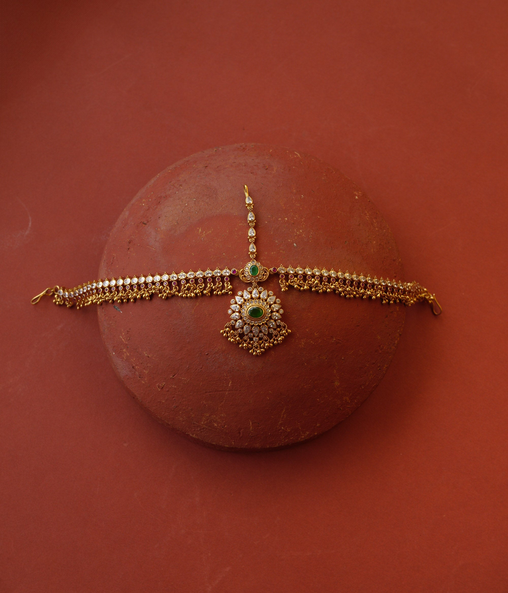 Traditional gold mathapatti with green spinal stone and Swarovski crystals in peacock eye-shaped central design.
