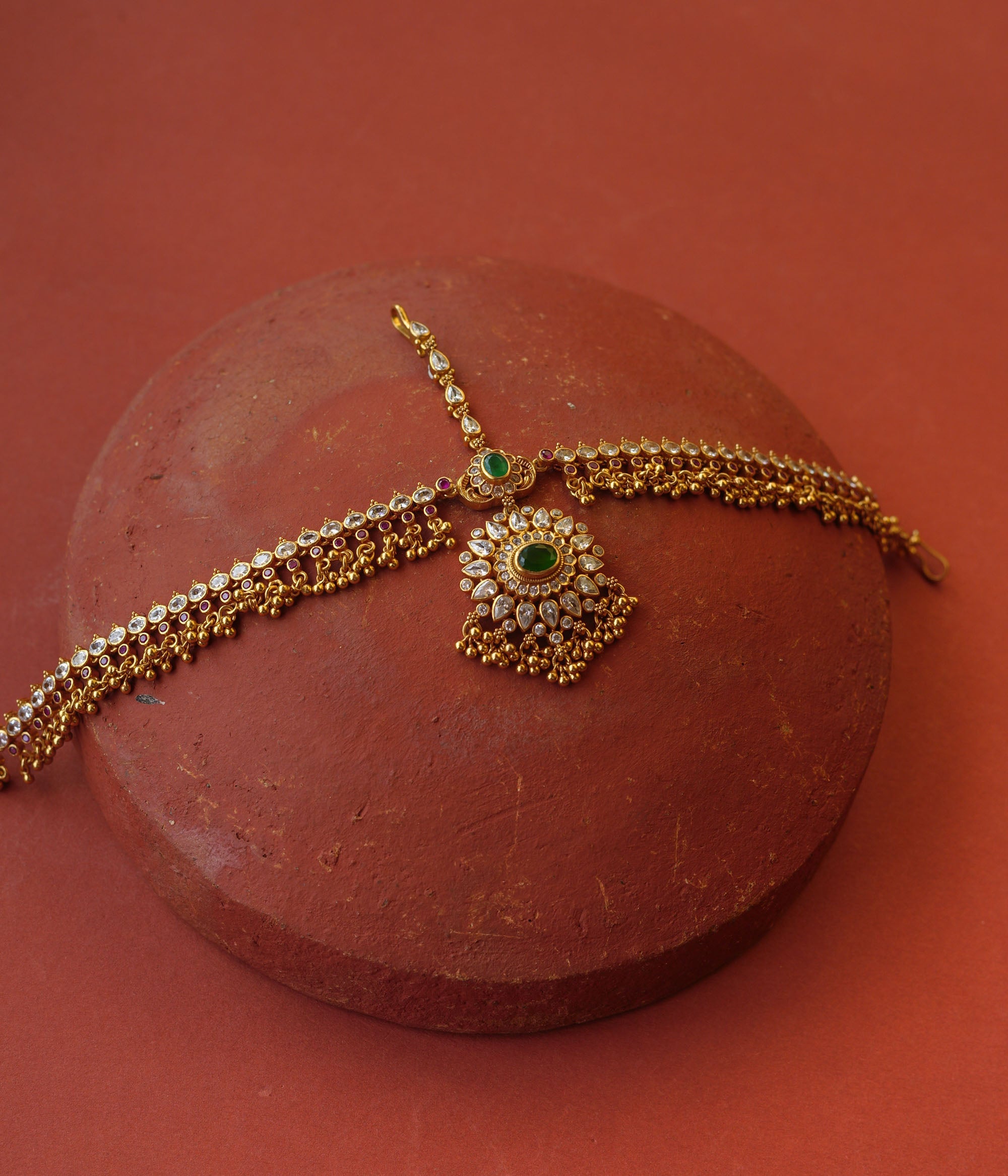 Traditional gold mathapatti with green spinal stone and Swarovski crystals in peacock eye-shaped central design.
