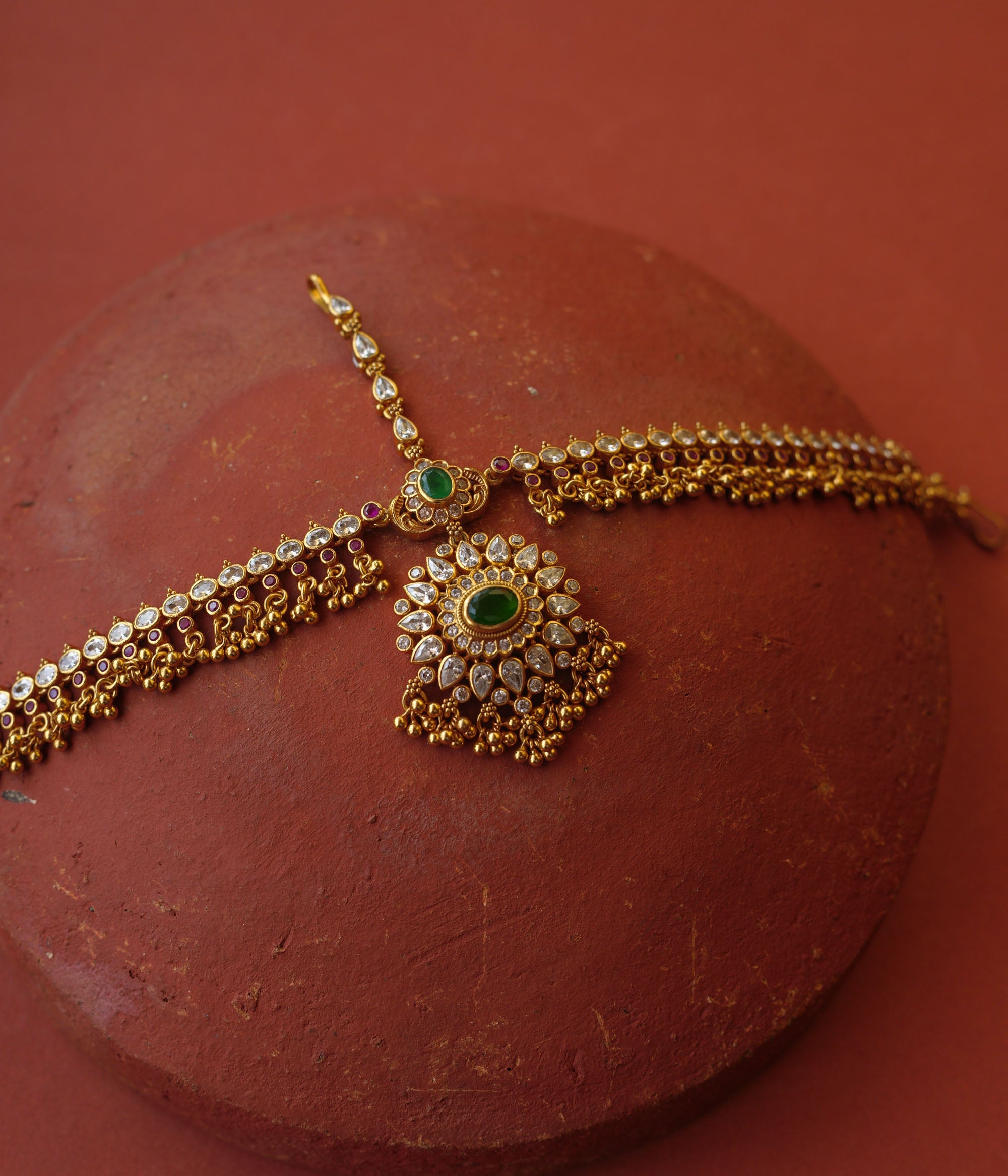 Traditional gold mathapatti with green spinal stone and Swarovski crystals in peacock eye-shaped central design.