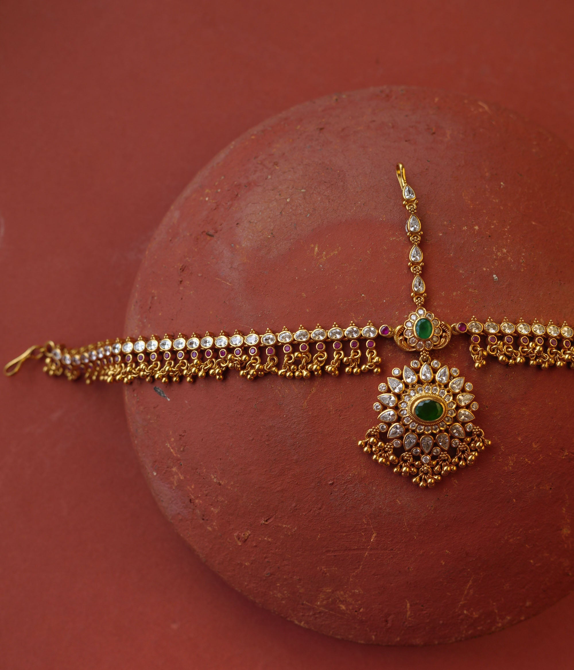 Traditional gold mathapatti with green spinal stone and Swarovski crystals in peacock eye-shaped central design.