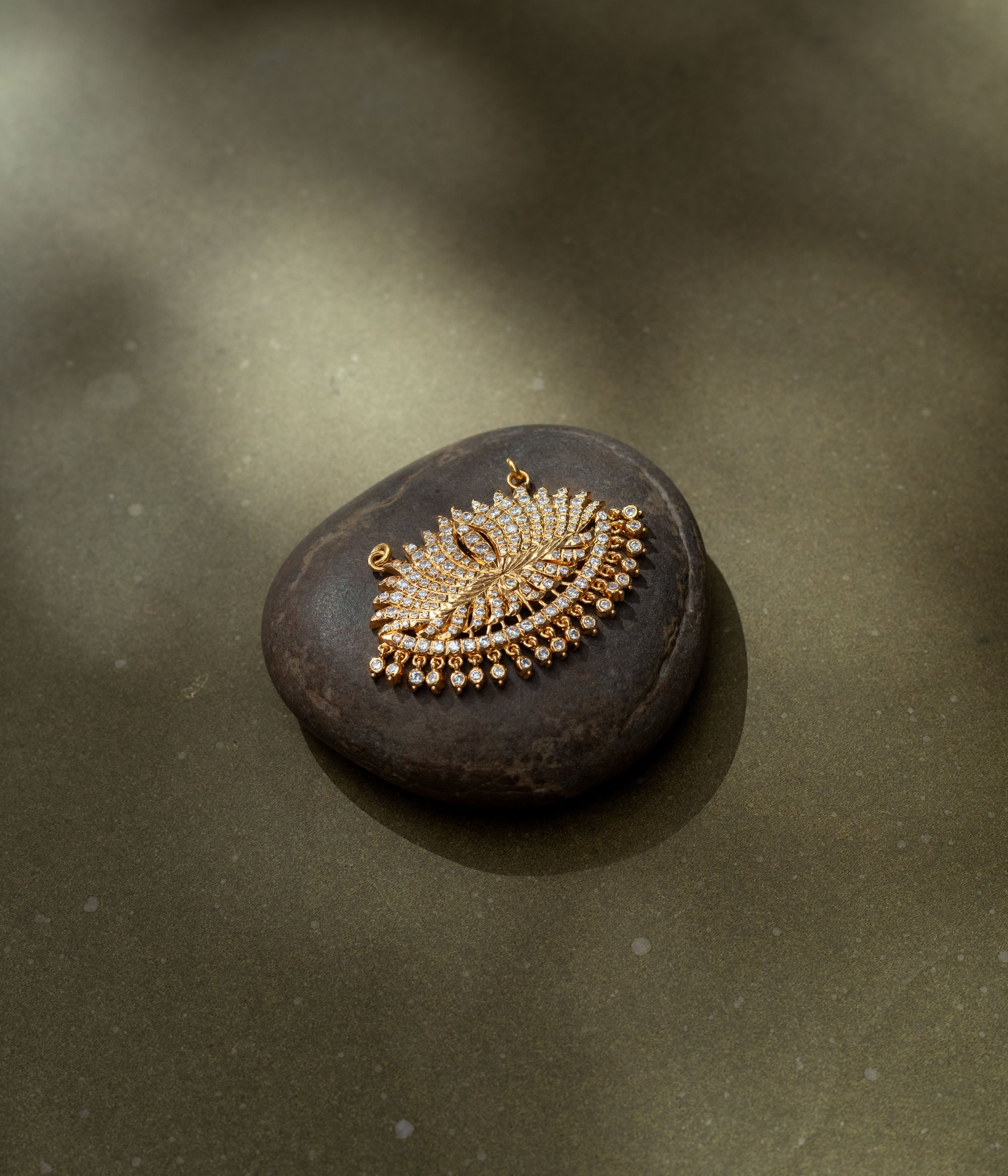 Gold fan-shaped pendant with CZ stones, inspired by peacock feather and sunburst motifs.
