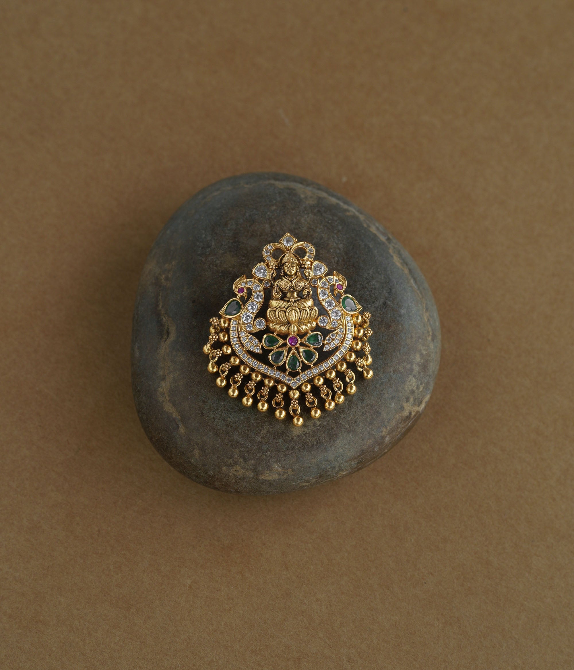 A gold pendant featuring a central deity figure on a lotus. It is decorated with clear, green, and red stones and has dangling gold beads.