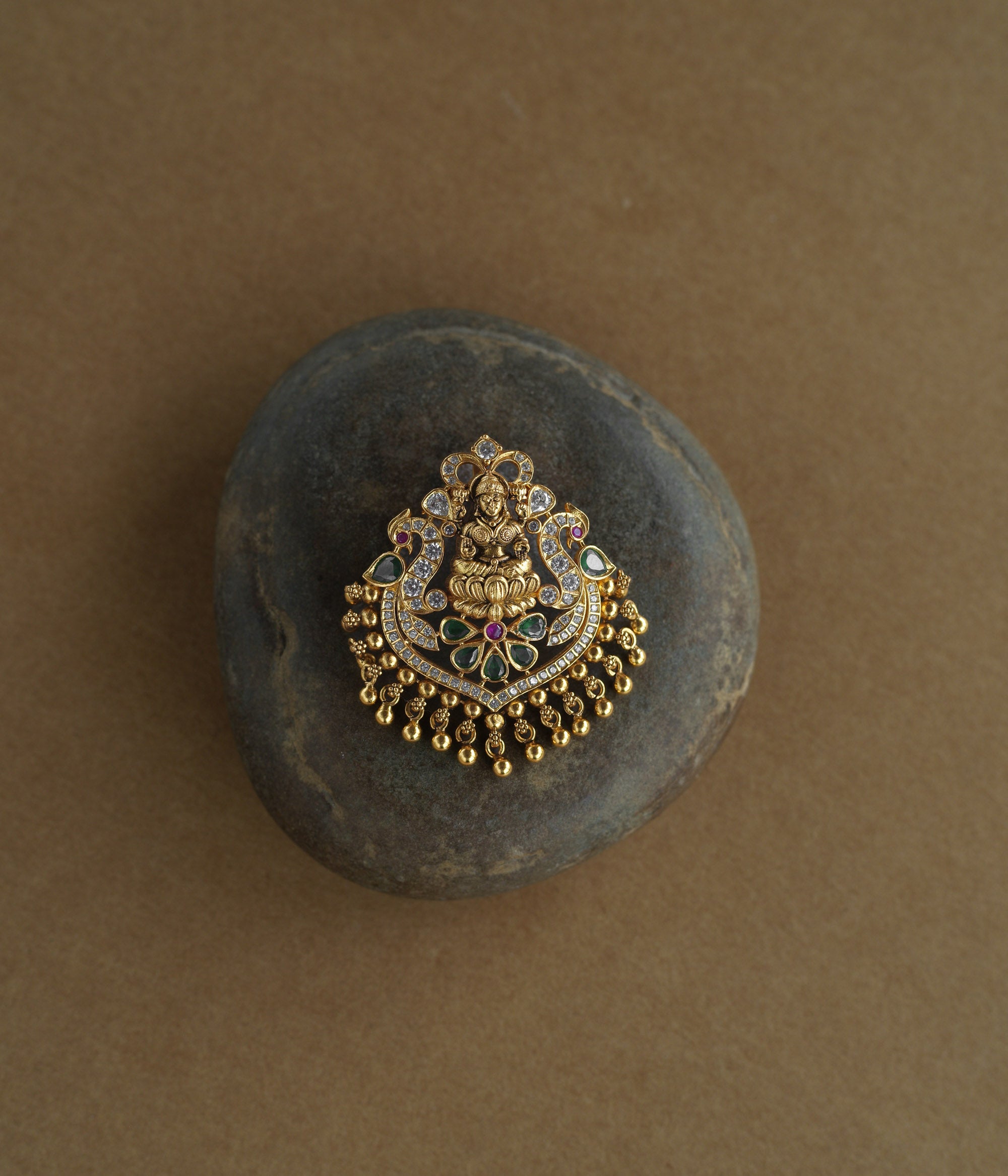 A gold pendant featuring a central deity figure on a lotus. It is decorated with clear, green, and red stones and has dangling gold beads.