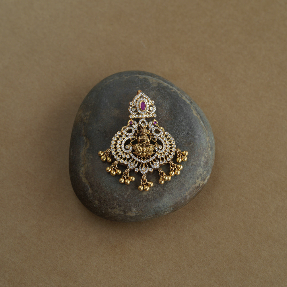 A gold pendant featuring a central deity figure. It is decorated with clear and red stones and has dangling gold beads.