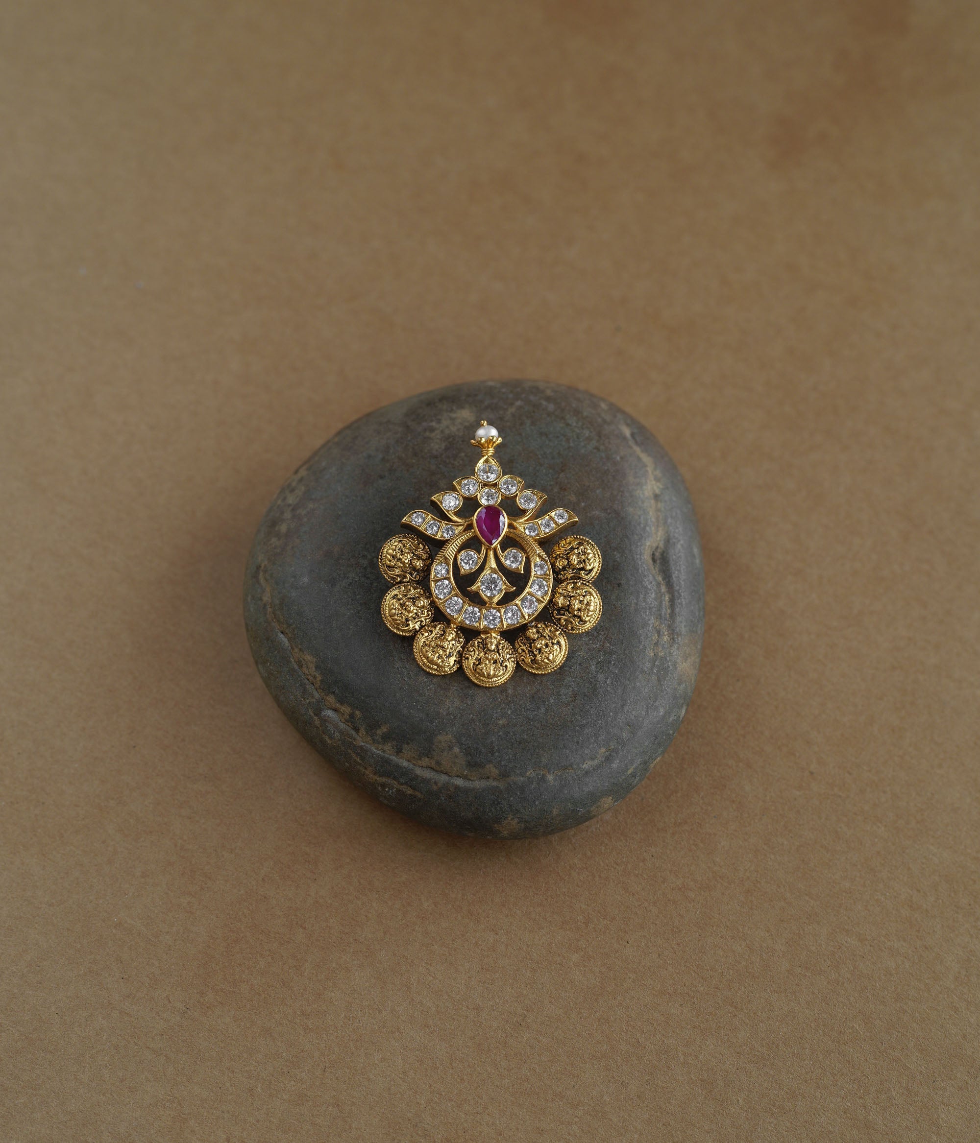 A golden pendant with a crescent-moon and floral design. It features a central red stone surrounded by clear stones and a halo of golden beads, resting on a stone.