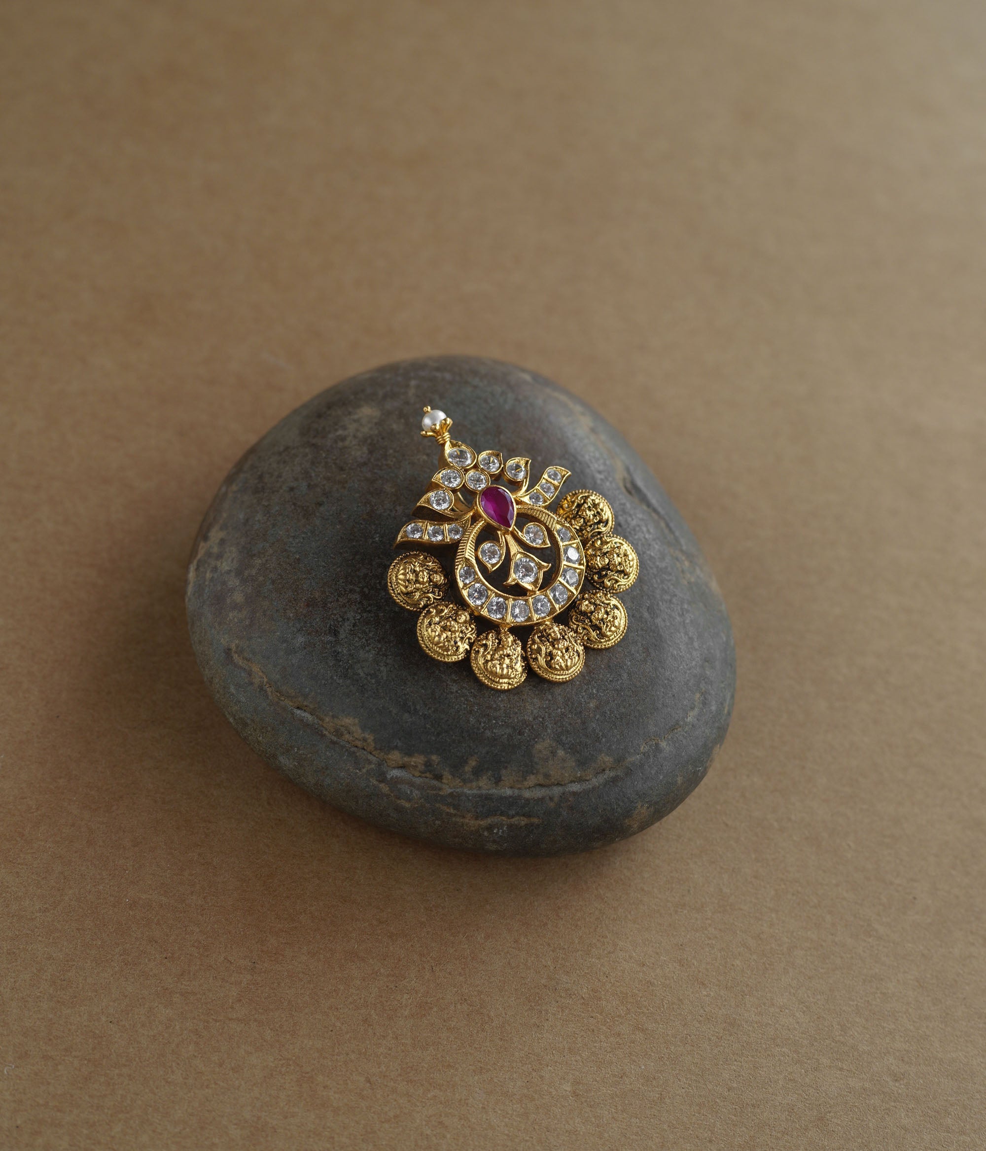 A golden pendant with a crescent-moon and floral design. It features a central red stone surrounded by clear stones and a halo of golden beads, resting on a stone.