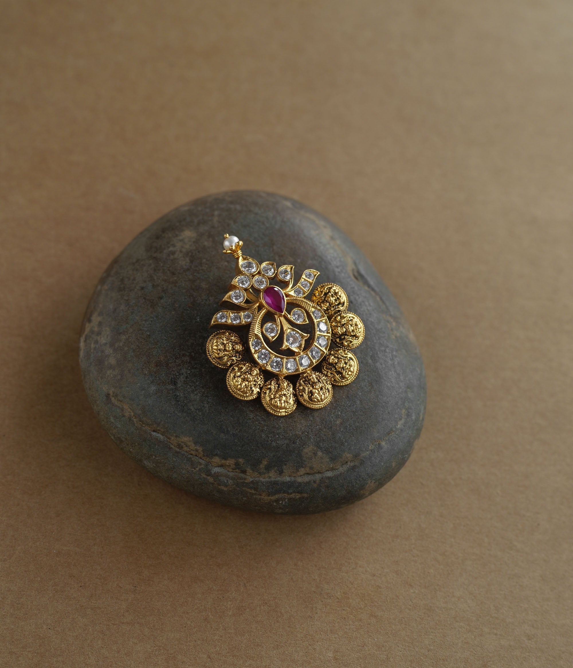 A golden pendant with a crescent-moon and floral design. It features a central red stone surrounded by clear stones and a halo of golden beads, resting on a stone.