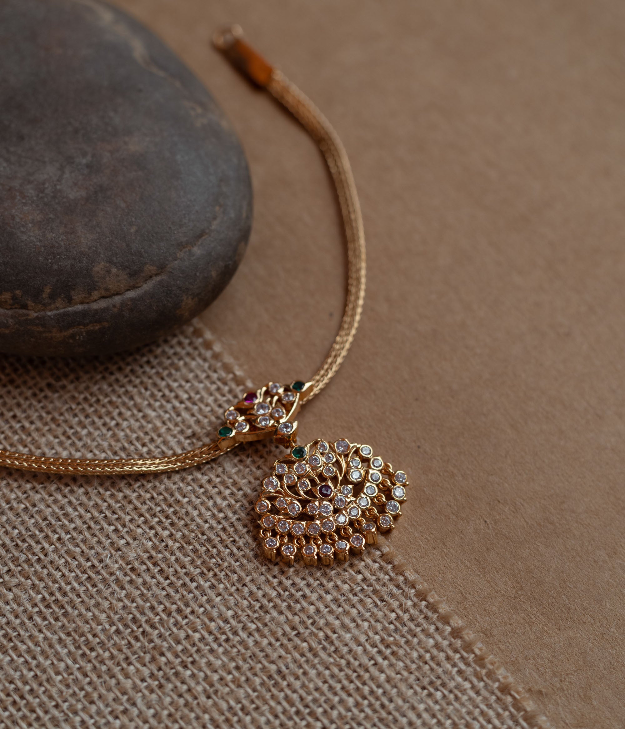 Close-up of a gold necklace with a tree-shaped pendant. The pendant is covered in clear, red, and green stones, attached to a woven gold chain.