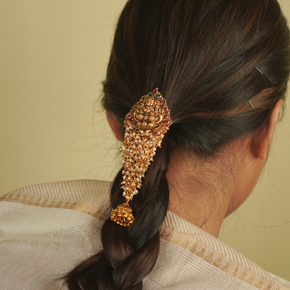 Saesha Hairpin: A gold-plated temple motif adorned with cascading pearls, red and green spinel stones, and a delicate jhumki. A graceful fusion of tradition and elegance for your hair.