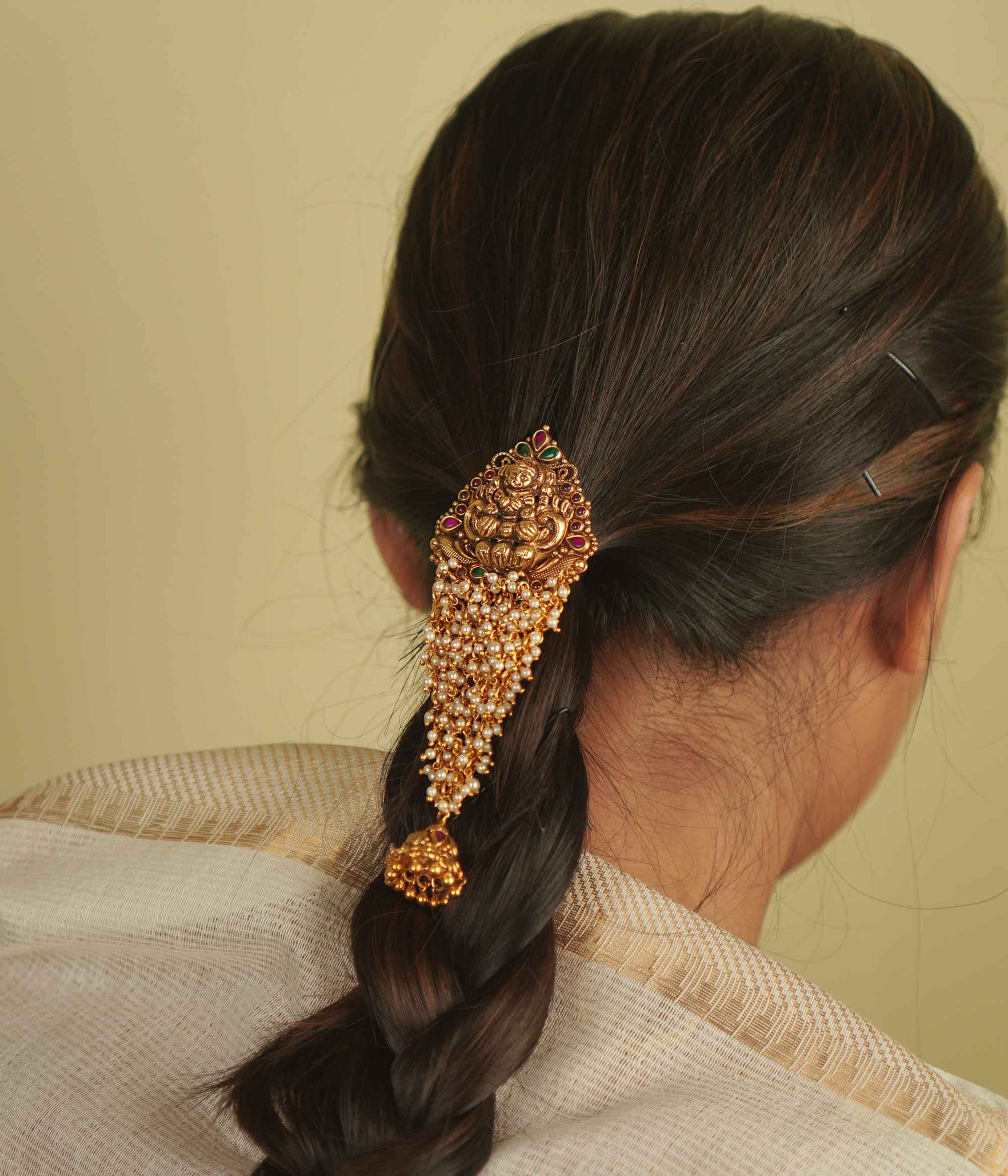Saesha Hairpin: A gold-plated temple motif adorned with cascading pearls, red and green spinel stones, and a delicate jhumki. A graceful fusion of tradition and elegance for your hair.