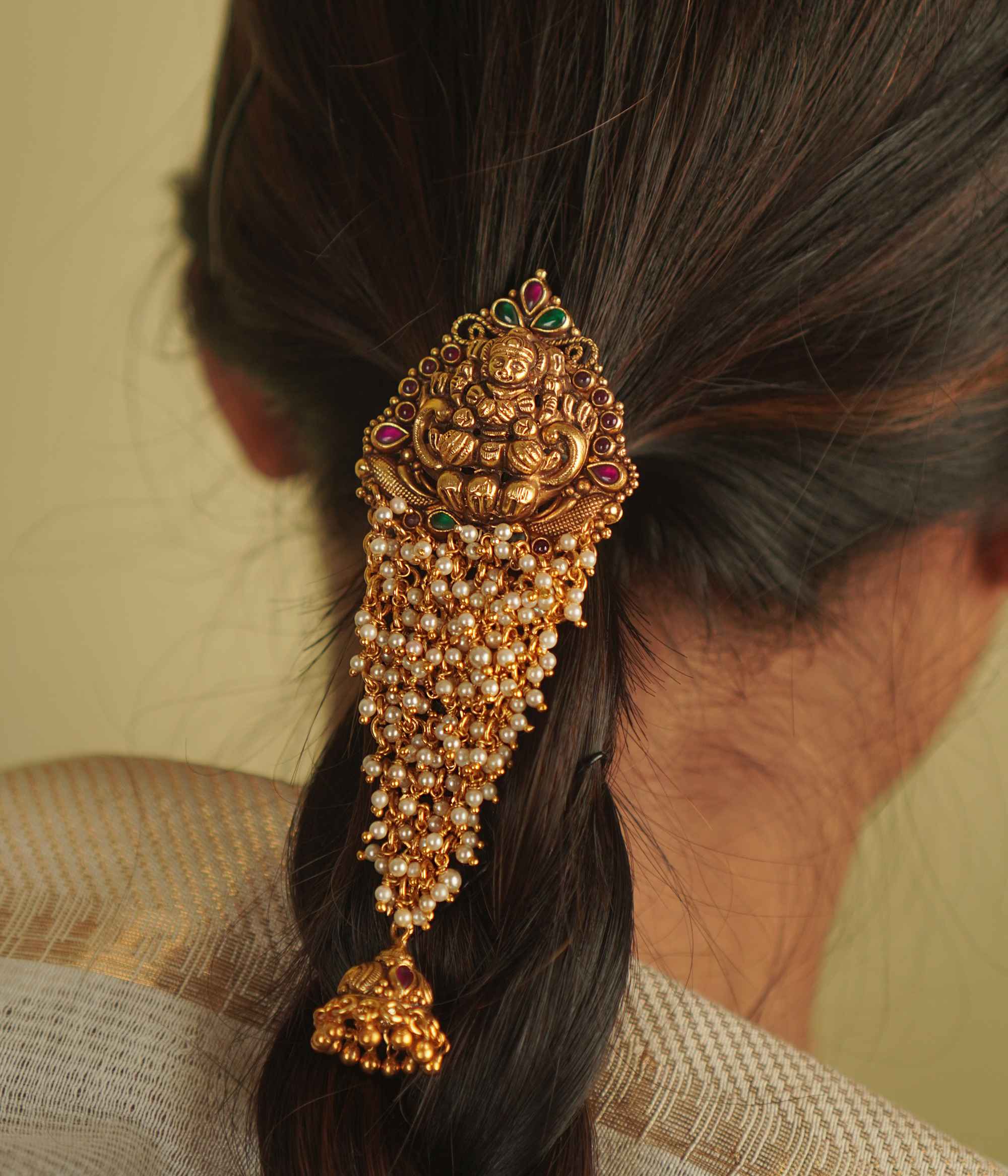 Saesha Hairpin: A gold-plated temple motif adorned with cascading pearls, red and green spinel stones, and a delicate jhumki. A graceful fusion of tradition and elegance for your hair.