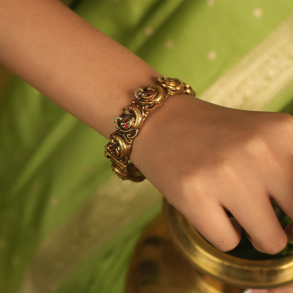 Embrace royal elegance with this stunning antique gold bangle, adorned with vibrant red, white, and green Kundan stones, featuring an intricate peacock motif. The regal peacock comes to life in this piece, while the pink Kundan hues add a touch of grandeur. A perfect fusion of heritage and artistry, this bangle exudes timeless splendor.
