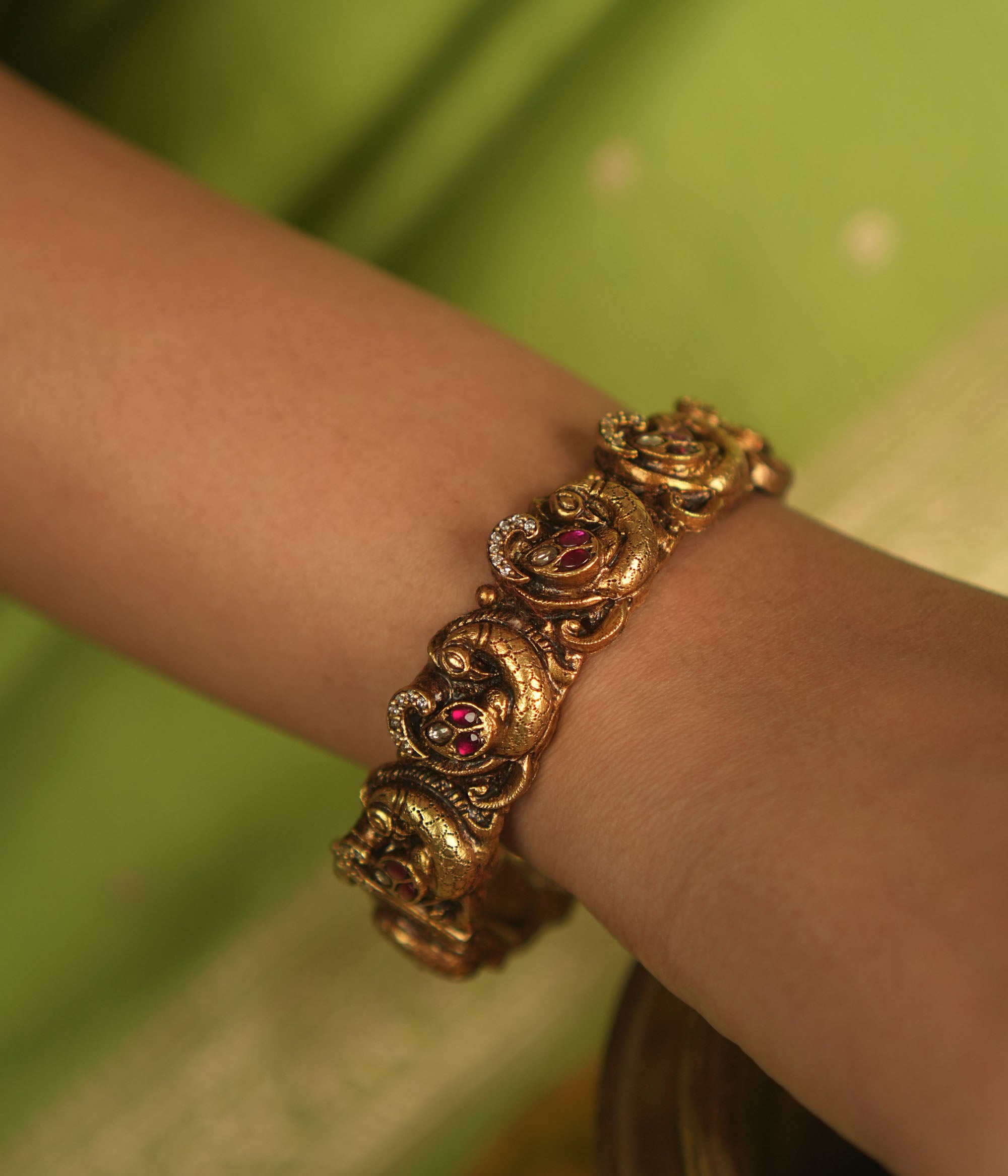 Embrace royal elegance with this stunning antique gold bangle, adorned with vibrant red, white, and green Kundan stones, featuring an intricate peacock motif. The regal peacock comes to life in this piece, while the pink Kundan hues add a touch of grandeur. A perfect fusion of heritage and artistry, this bangle exudes timeless splendor.
