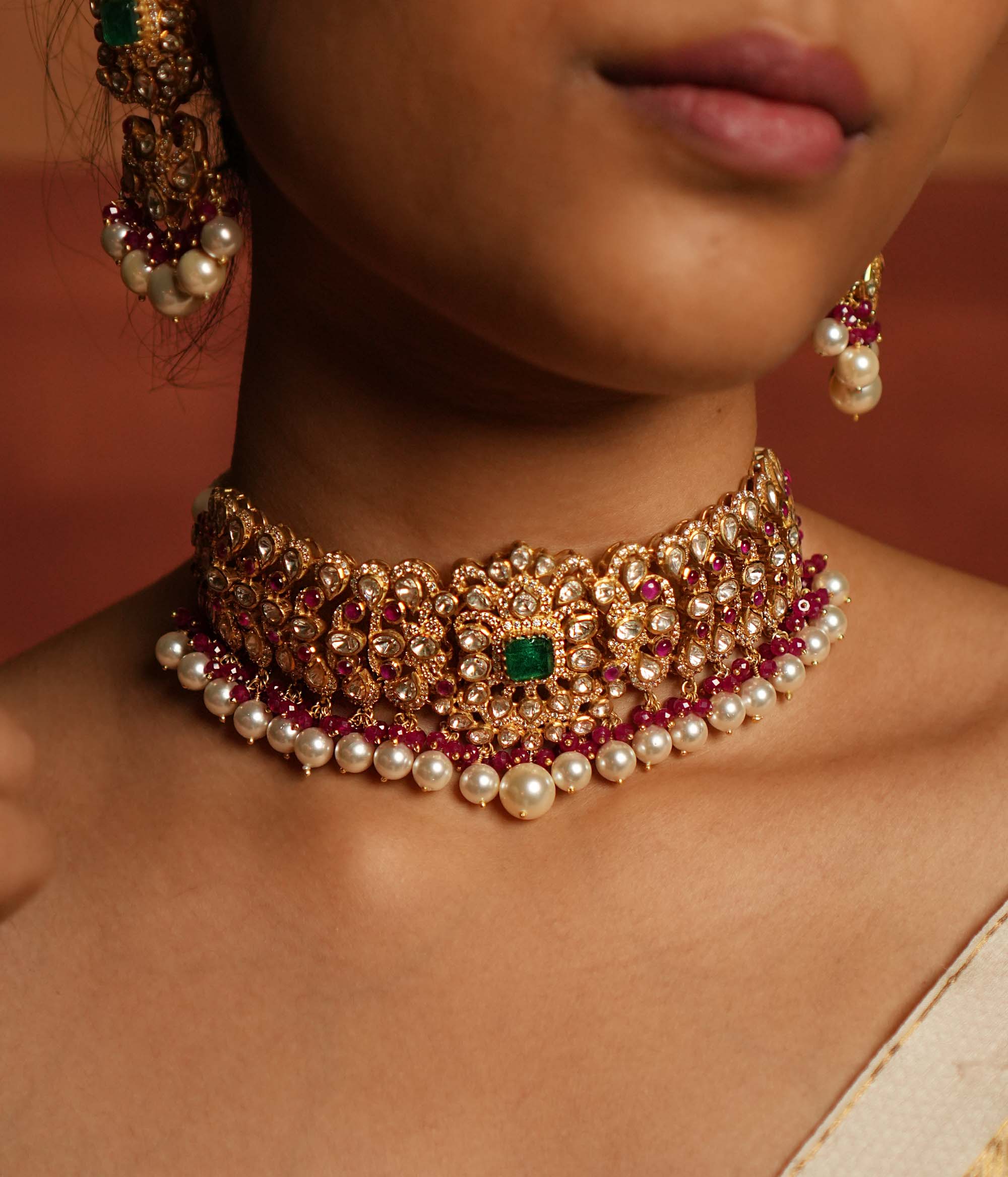 Exquisite choker featuring a central floral design adorned with Green Gemstone, Moissanite Polki Stones, Red Beads, Pearls, and CZ Stones. The choker showcases a continuous chain of Polki and Red Beads, elegantly finished with delicate pearl drops for a regal look.
