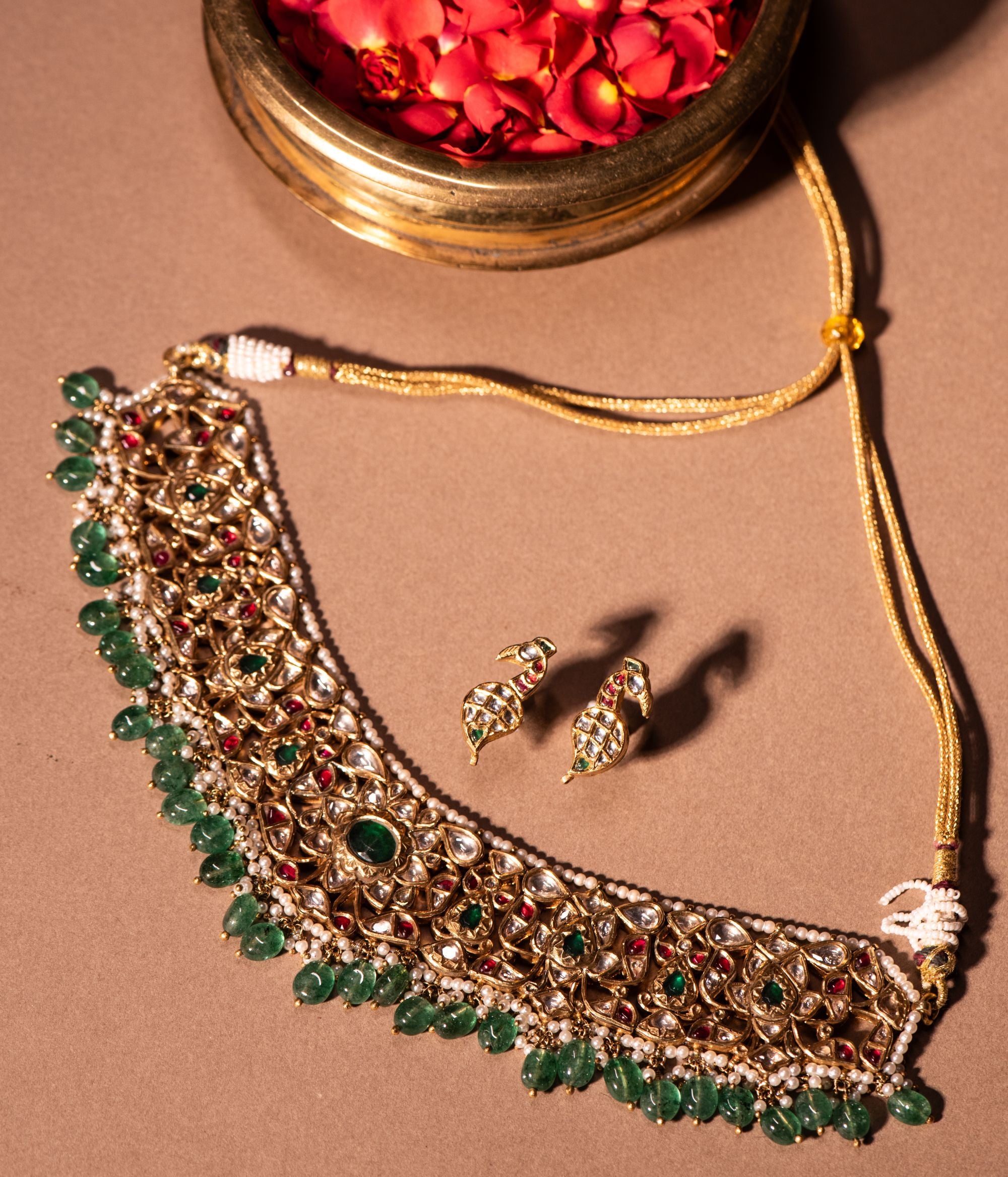 An opulent gold-plated choker necklace adorned with sparkling white sapphires, vibrant rubies, rich emeralds, and delicate pearls. The intricate lattice design is embellished with a striking combination of gemstones, while the emerald bead fringe adds a touch of regal elegance. Accompanied by matching earrings featuring a graceful peacock-inspired motif, this set exudes timeless charm and sophistication.