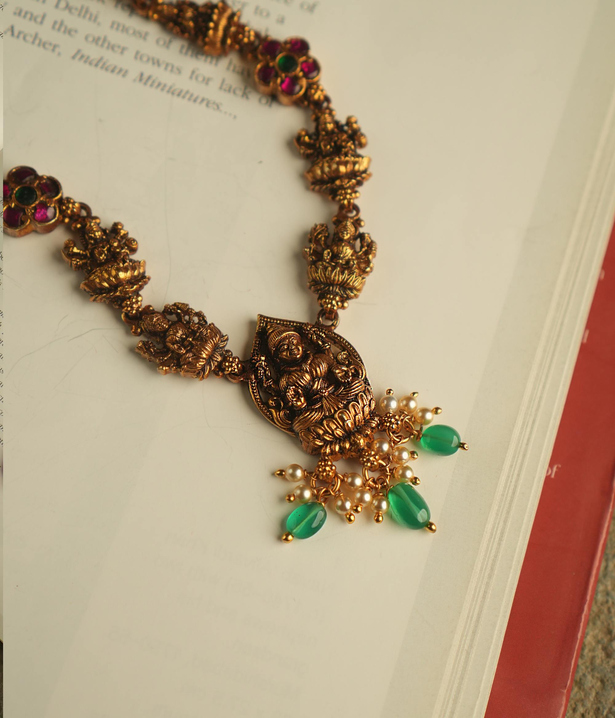 The Aadhya necklace evokes the charm of a bygone era, with antique gold, polkis, and a divine Lakshmi pendant. Pearl drops and green beads add a touch of timeless grace.