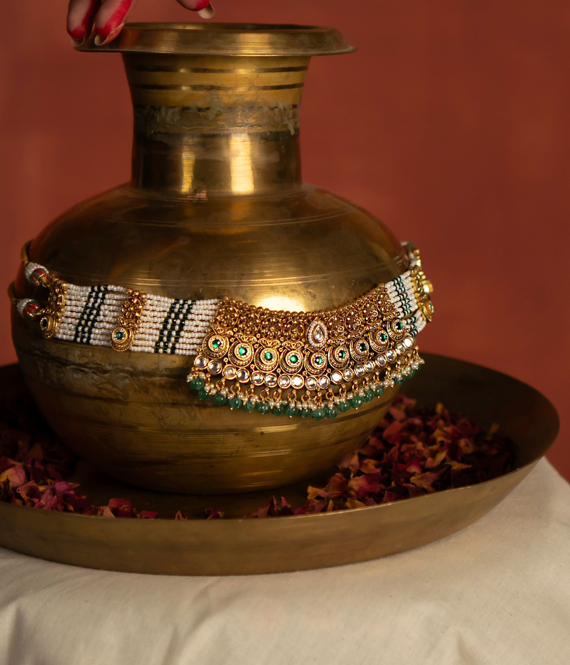 A gold necklace, richly embellished with white, green, and gold accents, is artfully draped over a vintage-style brass pot. The ornate necklace, with its intricate detailing, contrasts beautifully with the rustic texture of the pot. This juxtaposition highlights the craftsmanship of the necklace and creates a visually compelling composition.