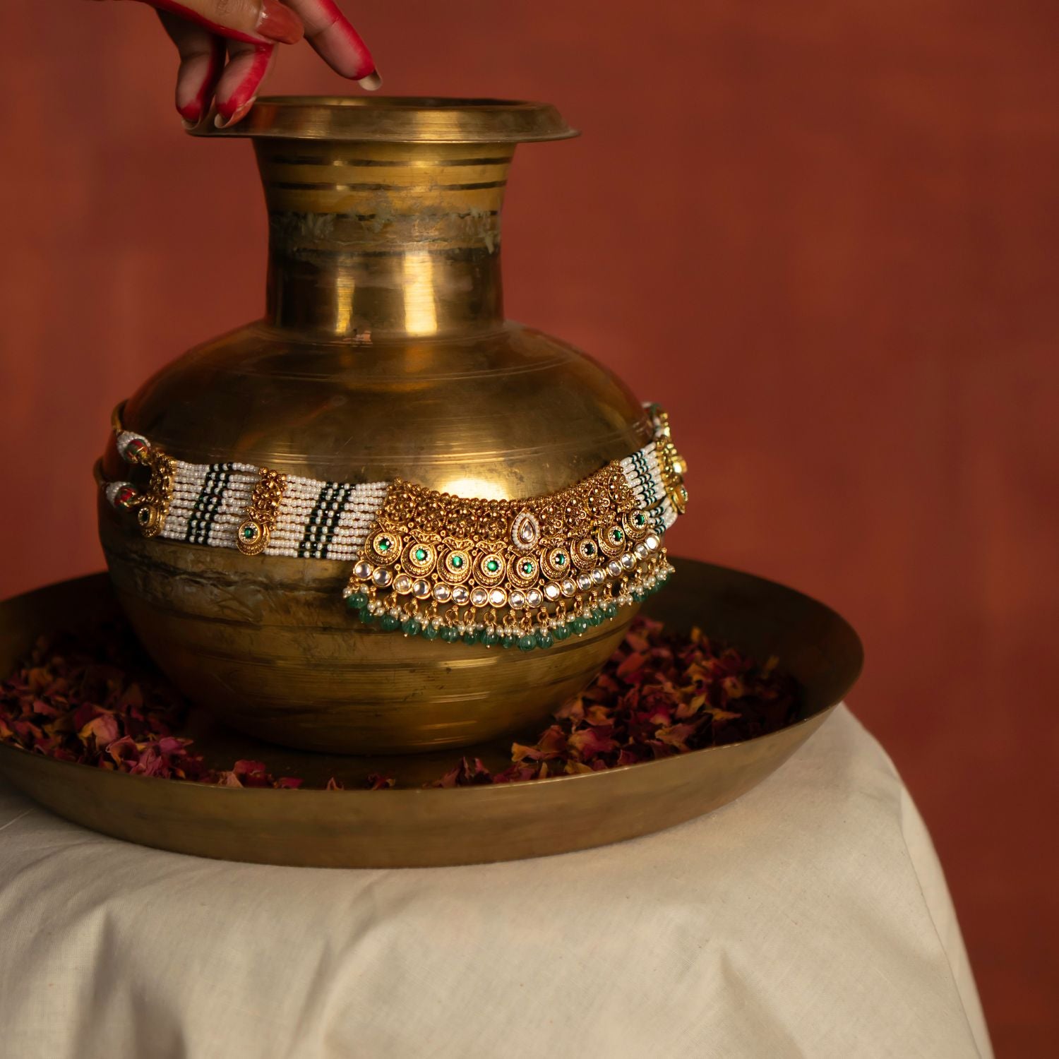 A gold necklace, richly embellished with white, green, and gold accents, is artfully draped over a vintage-style brass pot. The ornate necklace, with its intricate detailing, contrasts beautifully with the rustic texture of the pot. This juxtaposition highlights the craftsmanship of the necklace and creates a visually compelling composition.