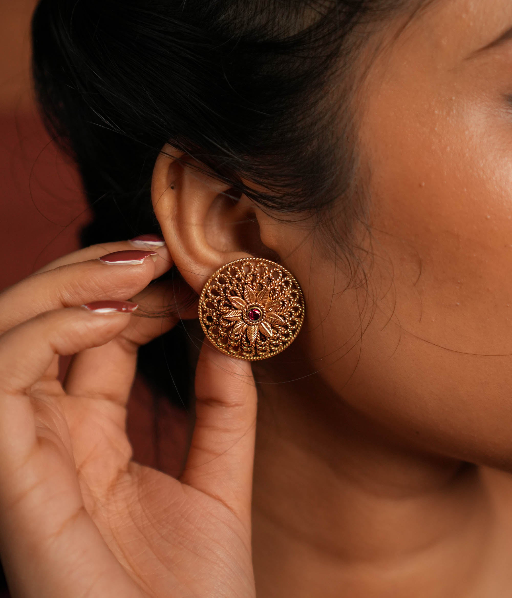 Round gold-plated silver earrings featuring a bold floral center with intricate petals and delicate lines, creating a harmonious design. Inspired by vintage Kolhapuri tribal artistry, the earrings showcase a radiant red gemstone heart at the center, adding a touch of timeless elegance.