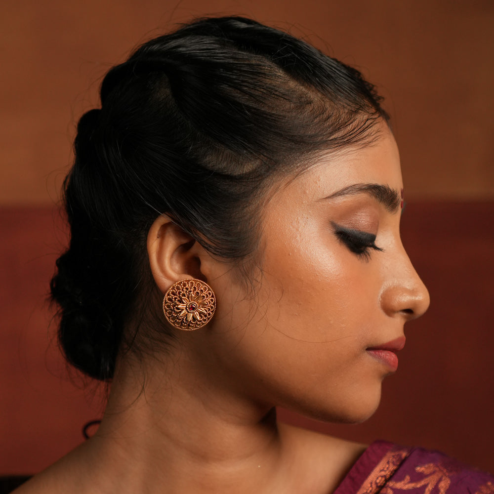 Round gold-plated silver earrings featuring a bold floral center with intricate petals and delicate lines, creating a harmonious design. Inspired by vintage Kolhapuri tribal artistry, the earrings showcase a radiant red gemstone heart at the center, adding a touch of timeless elegance.