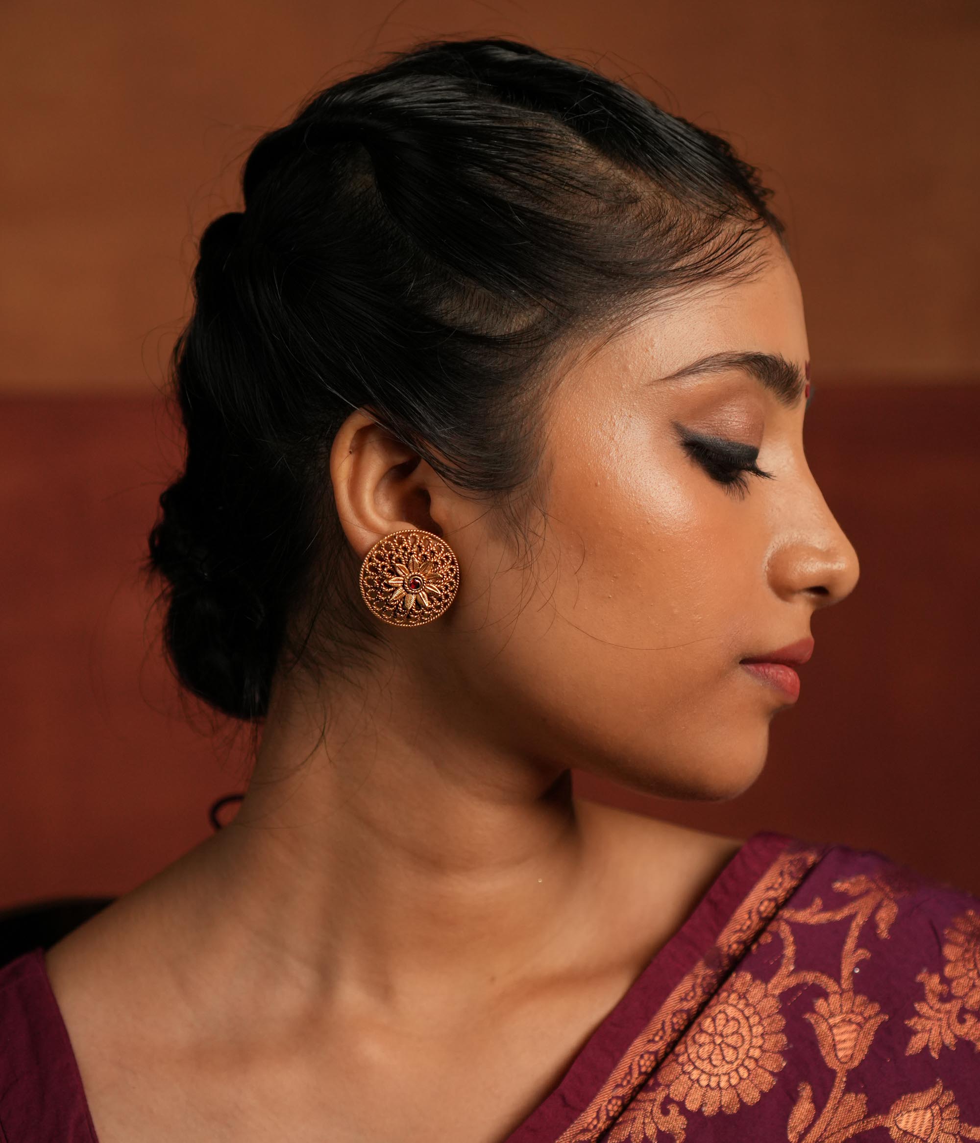 Round gold-plated silver earrings featuring a bold floral center with intricate petals and delicate lines, creating a harmonious design. Inspired by vintage Kolhapuri tribal artistry, the earrings showcase a radiant red gemstone heart at the center, adding a touch of timeless elegance.