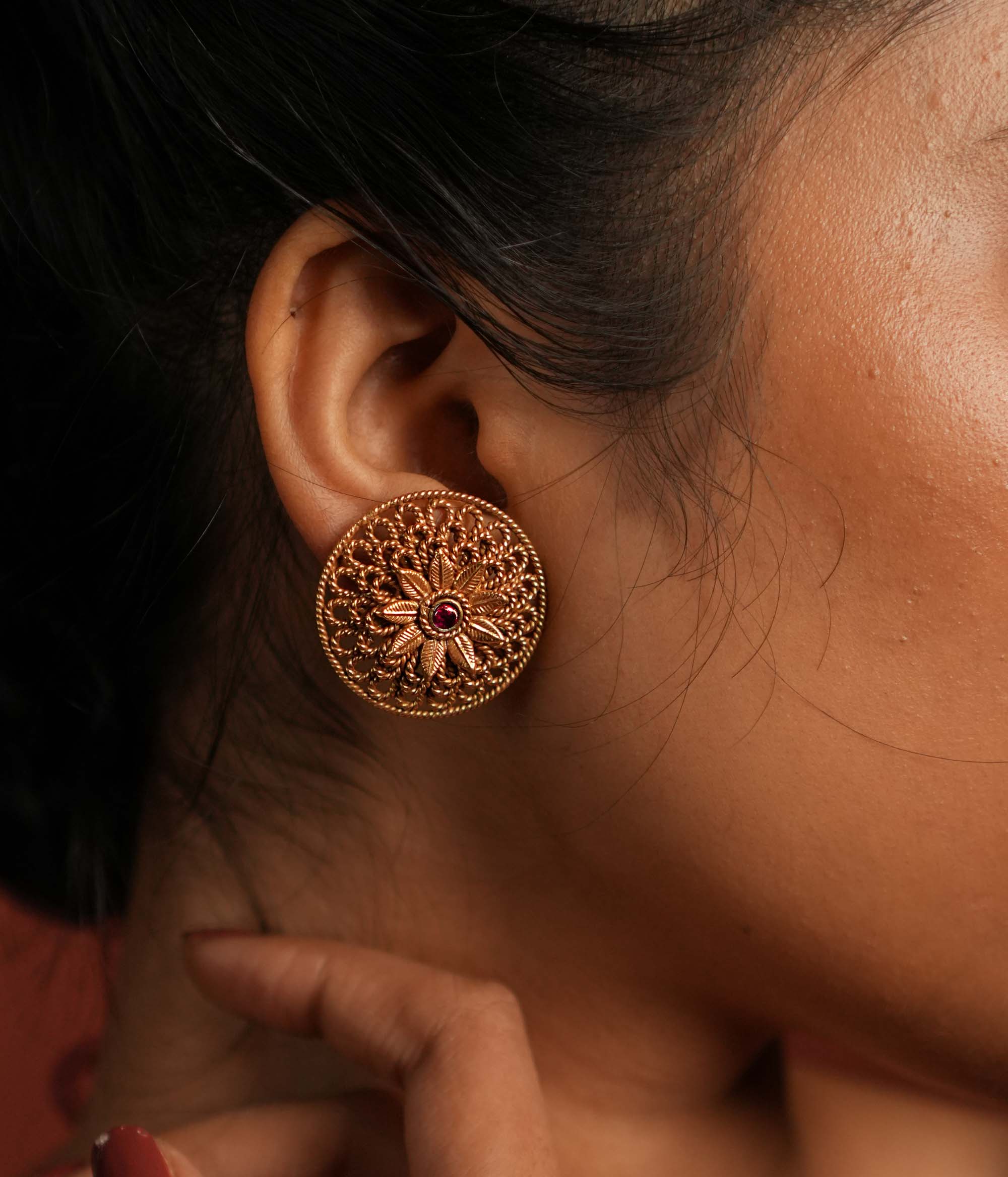 Round gold-plated silver earrings featuring a bold floral center with intricate petals and delicate lines, creating a harmonious design. Inspired by vintage Kolhapuri tribal artistry, the earrings showcase a radiant red gemstone heart at the center, adding a touch of timeless elegance.