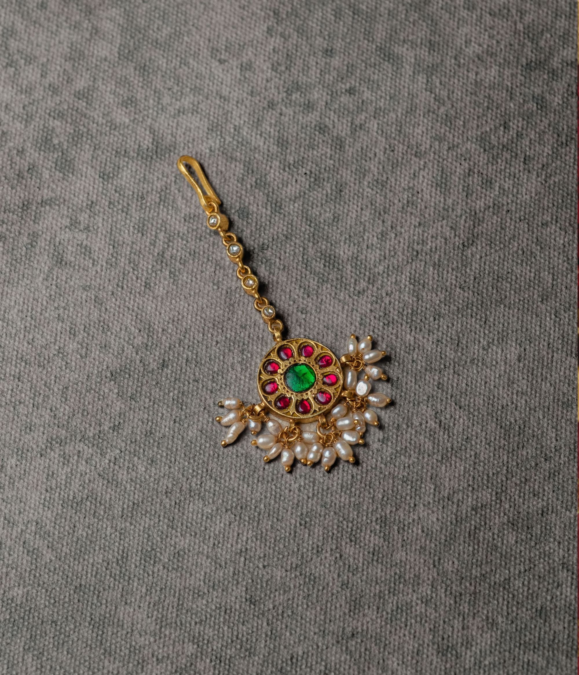 A cherished heirloom, this antique-inspired tikka features red and green Kundan stones with white pearls, embodying elegance and tradition through its timeless craftsmanship.