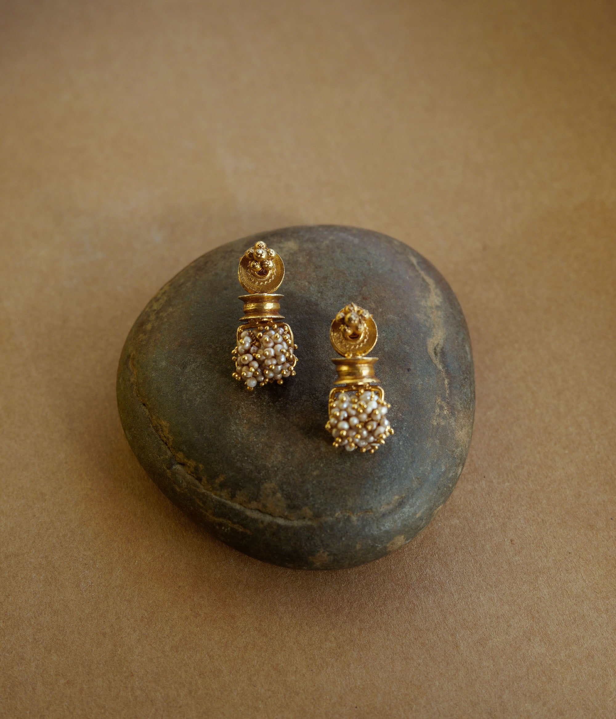 Gold earrings with a cylindrical pillar-like shape. A cluster of small white pearls hangs from the base. The earrings are on a textured brown surface.