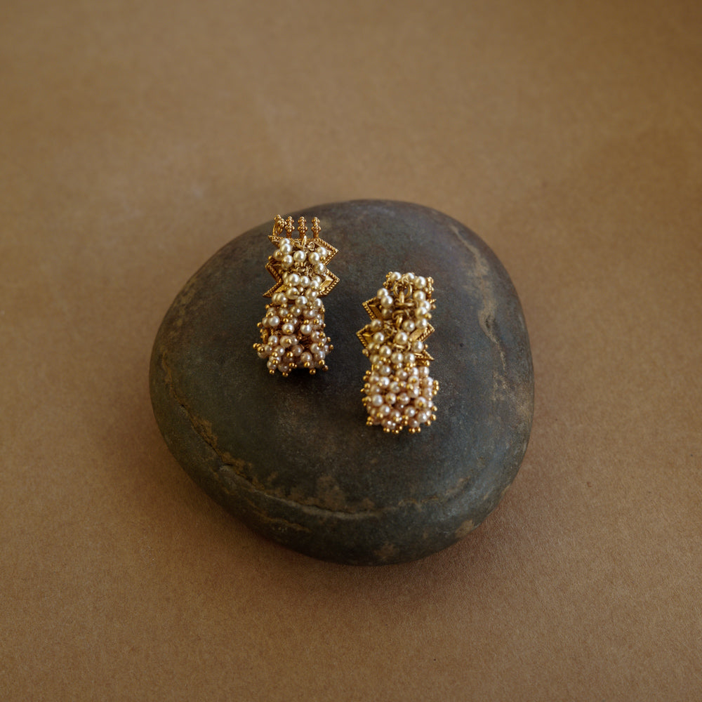 Gold earrings with a jagged, star-like motif at the top and a cluster of golden-hued beads/pearls dangling below. The earrings are on a textured brown surface.
